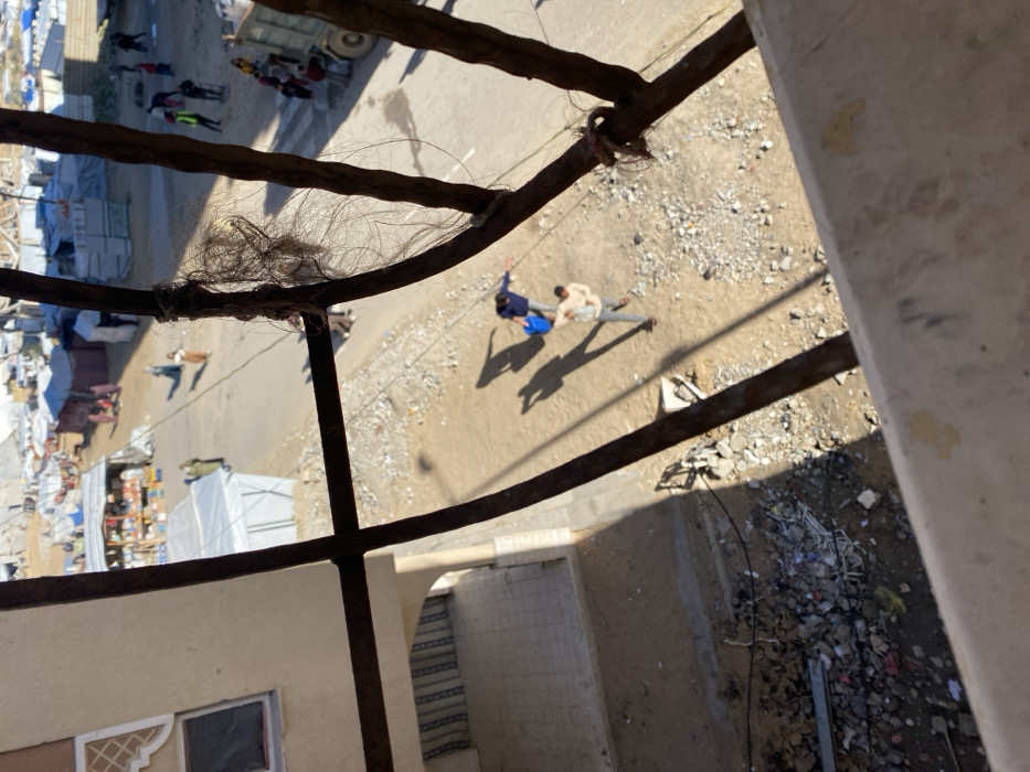 A high-angle shot, potentially from a window or balcony, shows a street scene. Two men are walking together, one in a blue shirt. A market area is visible in the distance, with white tents and people. The foreground includes an iron railing or bars, partially obscured by what appears to be tufts of hair or debris. The surrounding architecture is weathered and aged, with a nearby archway. The ground is a mix of pavement and loose debris.
