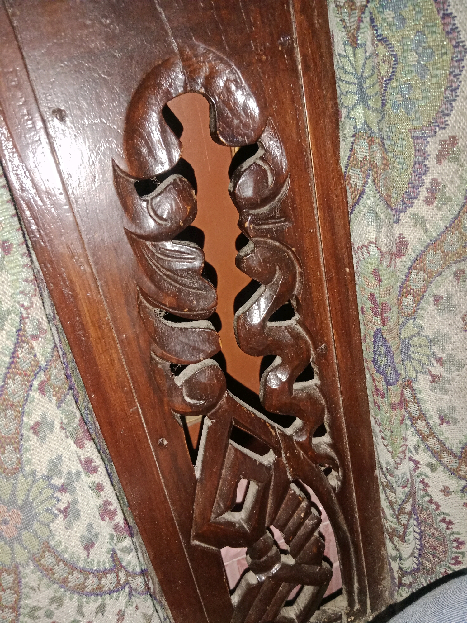 The image shows a close-up of intricately carved dark brown wood, likely part of a piece of furniture. The carving features an ornate, stylized pattern with curved lines and geometric shapes. A patterned fabric, with shades of green, purple, and beige, is visible behind and to the right of the wooden carving. The wood appears to be aged and shows some dust accumulation. The scene is indoors, with a neutral background visible through the carved openings. There are no people visible in the image, and no specific location in Ringinrejo, Indonesia can be identified.