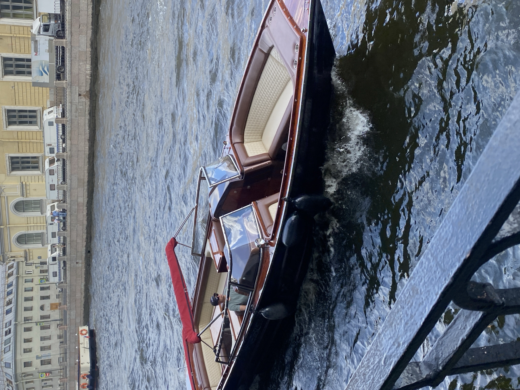 The image shows a man operating a small, wooden motorboat with a red canopy on a river in Saint Petersburg, Russia. The boat has luxurious brown and cream-colored seating. The man is wearing a green t-shirt and a black baseball cap, and sunglasses. The river has choppy water, indicating movement. In the background, across the river, there is a stone embankment lined with buildings and a walkway. A white truck with "Worldwide Logistics" printed on it and several white camper vans are visible on the walkway, suggesting a street or road adjacent to the embankment. There are also pedestrian railings and some trees. Another boat, a white passenger vessel with orange life preservers, is visible further down the river on the left. The overall scene depicts a typical day on a canal or river in Saint Petersburg.
