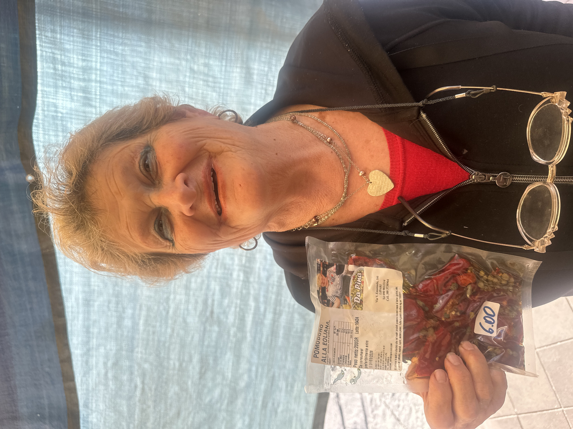 An older woman smiles warmly, looking directly at the viewer while holding a clear plastic bag of food items. She has light brown hair, is wearing a black zippered jacket over a red top, and a silver necklace with a heart-shaped pendant. Her eyeglasses hang from a cord around her neck.
The bag she holds contains dried red items, identified by the label as "POMODORO ALLA EOLIANA" (Aeolian style tomatoes) and also mentions "capperi" (capers). A sticker on the bag displays the price "6.00" (likely Euros). The label also provides detailed information: "Da Pina" as the producer, located at "Via V. Emanuele, 166, LIPARI", along with phone numbers. Other visible text includes "Peso netto: 200GR", "Lotto: 16424", and a "De consumarsi preferibilmente entro 31/03/2028" date. An allergy warning is present, indicating possible traces of gluten, milk, peanuts, lupins, celery, fish, and tree nuts.
The background consists of a light blue, textured fabric, suggesting an outdoor market stall or a covered vendor area, consistent with the location of Lipari, Italy. The bright, diffused lighting indicates it is daytime. The woman's expression suggests she is a friendly vendor or presenter of the local product.