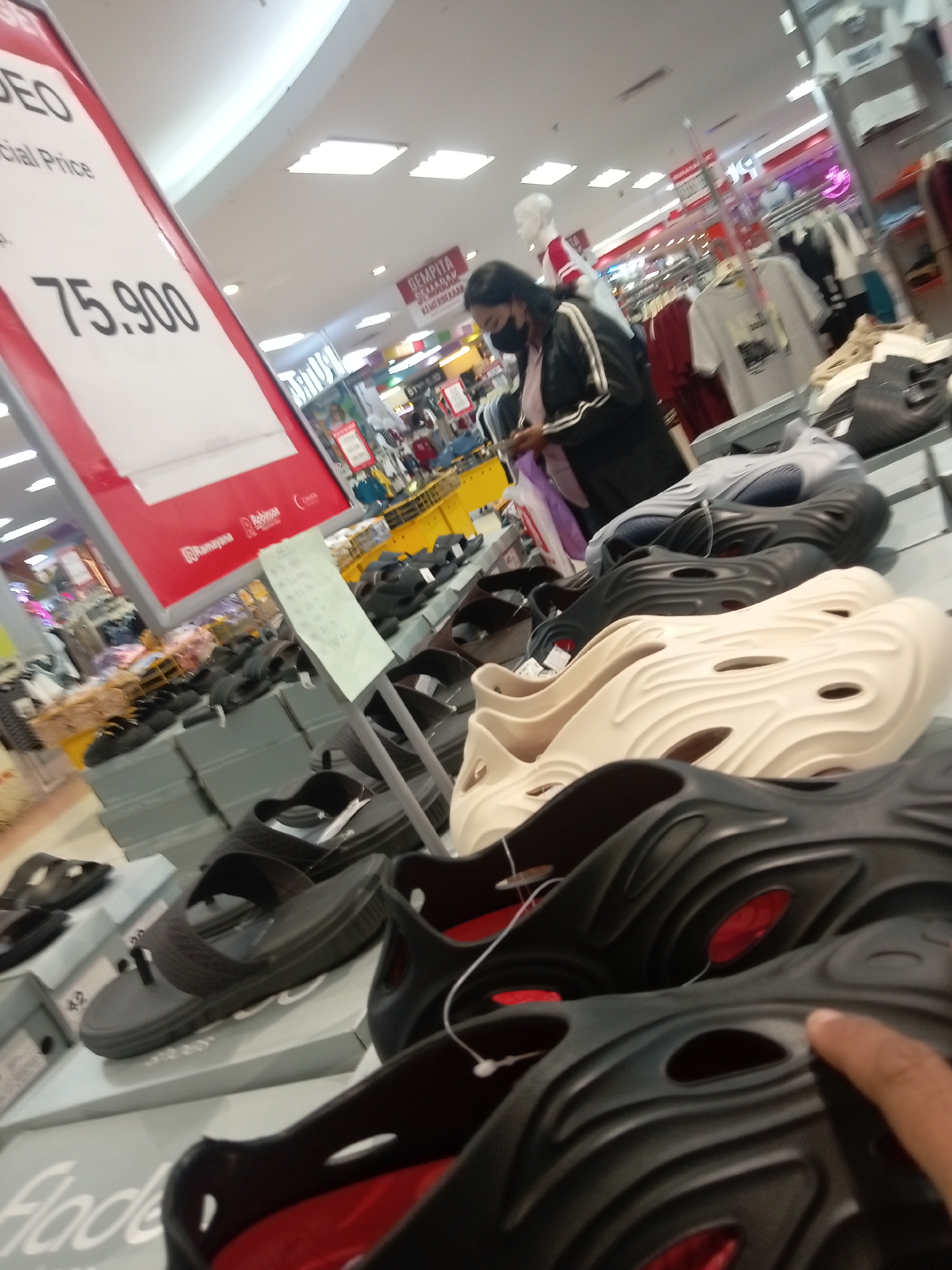 The image shows a display of sandals and shoes in a retail store. A price tag displays "75.900". In the background, a woman wearing a mask and a jacket is looking at her phone. Clothing racks are visible behind her, suggesting this is a department store. The store appears to be well-lit with fluorescent ceiling lights. The overall environment suggests a typical indoor shopping mall setting. Based on the price format and the likely context of a retail store in Indonesia, the location is likely a mall in Batam, Indonesia.