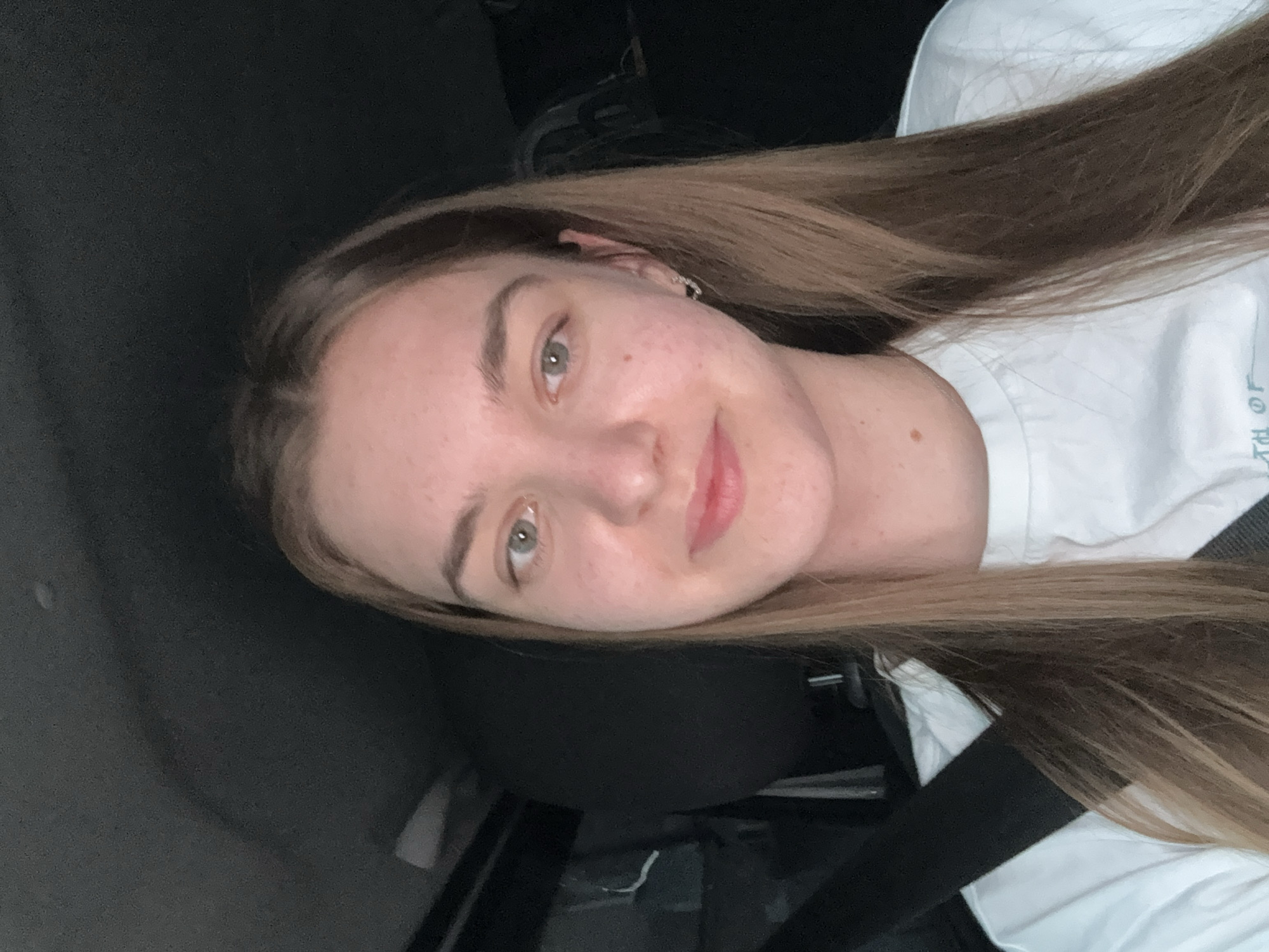 A young woman with light brown hair and fair skin is positioned in what appears to be the front seat of a car. She is wearing a white shirt and a black seatbelt is visible across her chest. The interior of the car is dark, suggesting low lighting. Her facial expression is calm with a slight smile. The exact location in Perm, Russia is not discernible from the image.