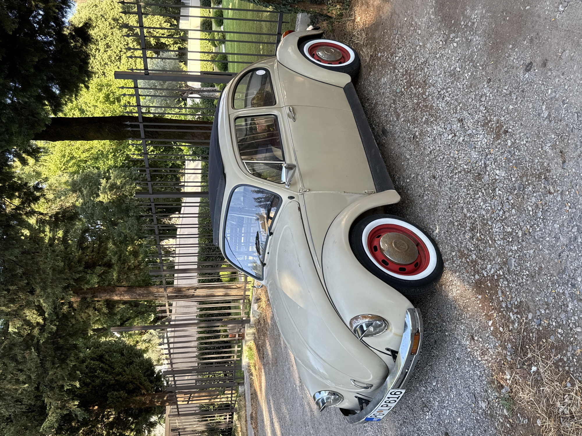 A vintage, beige Volkswagen Beetle is parked on a gravel lot outdoors. The car has a black sunroof and distinctive red and white hubcaps. Behind the car, a black metal fence separates the lot from a manicured green lawn and trees. The lighting suggests it is daytime, with sunlight casting shadows on the ground. The license plate of the car is visible and reads "PG LP616".