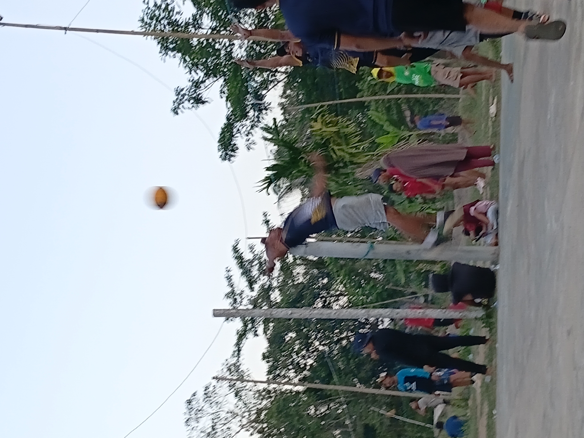 A group of people are playing volleyball outdoors. A male player in a blue and yellow shirt is in mid-air, about to hit the ball. Another male player in a black shirt and a blue hat is watching. Other spectators are visible in the background. The game is taking place on a dirt court with trees and foliage surrounding it. A volleyball net is strung between two poles. The sky is clear and overcast. The scene appears to be in Margo Rukun, Indonesia.