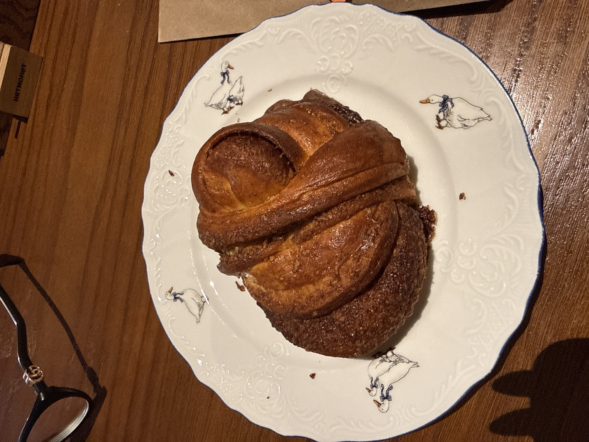 A close-up, eye-level shot captures a single, golden-brown, swirled pastry resting on a white, scalloped-edge plate adorned with small, hand-drawn duck motifs. The pastry appears to be a cinnamon roll or similar sweet bread, with a glossy sheen and visible layers of dough. The plate is set on a dark, wooden surface, likely a table. In the upper left corner, a pair of black-rimmed glasses with a gold "GG" logo on the temple is partially visible, casting a shadow. In the upper right, a small, brown cardboard sign with black text reading "НЕТ МОНЕТ" (meaning "No Coins" in Russian) is propped up. The overall scene suggests a casual setting, possibly a cafe or restaurant. The provided image does not contain any specific visual cues that would allow for the determination of its location within Moscow, Russia.