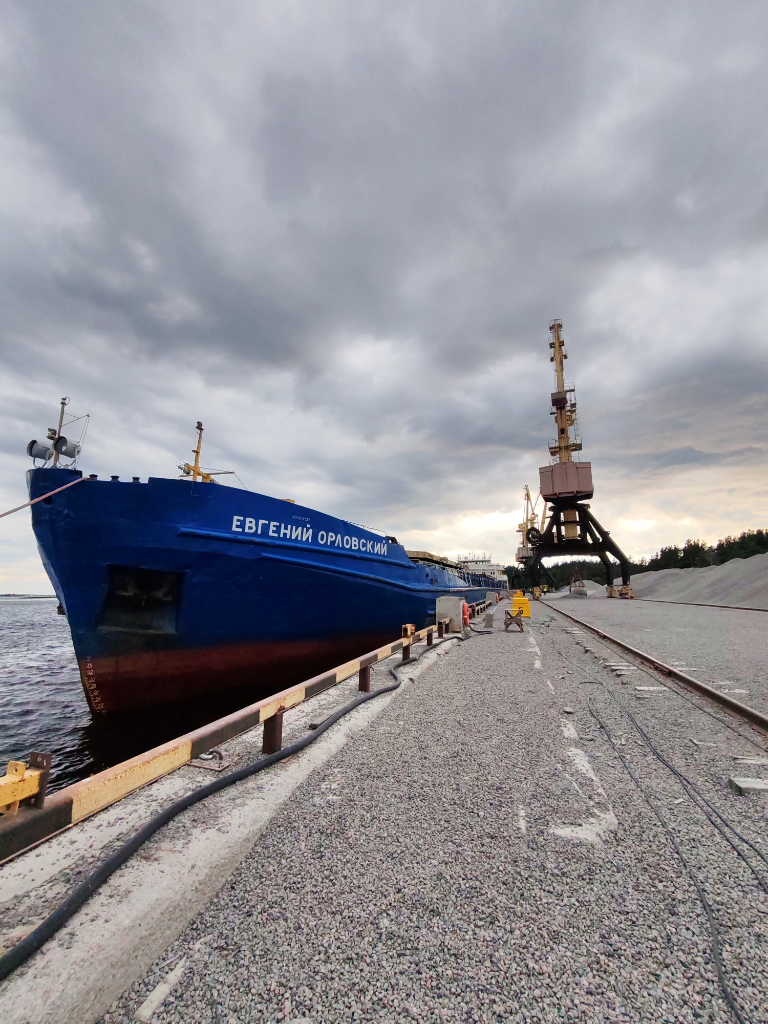 The image depicts a blue cargo ship named "Евгений Орловский" docked at a port in Shal'skii, Russia. The ship is positioned alongside a concrete pier covered in gravel. In the background, industrial cranes stand tall, indicating port operations. Several piles of gravel are visible on the right side of the image, suggesting the cargo being handled. The sky is overcast with dark, heavy clouds. A few individuals are present on the pier, possibly port workers. The scene conveys a sense of industrial activity in a cloudy, maritime environment.