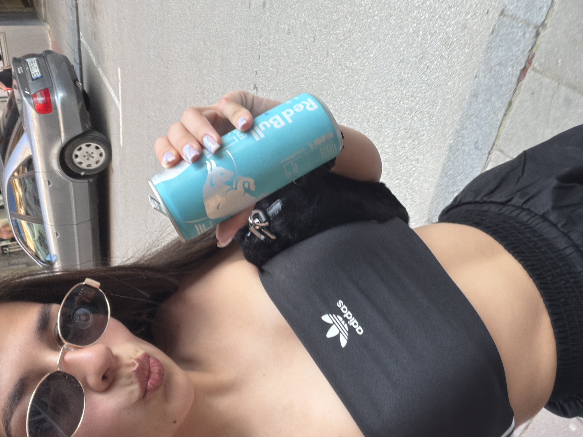 A young female is depicted in a close-up, waist-up outdoor shot in Gabrovo, Bulgaria. She is holding a light blue Red Bull energy drink can in her right hand, displaying light-colored fingernail polish with faint blue and pink marbling. She wears oval, gold-rimmed sunglasses, with a reflection of the can's pull-tab visible in the left lens (viewer's left). Her lips are pursed, covered in light pink lipstick. She wears a black strapless or bandeau crop top with a white Adidas Trefoil logo on the left chest. A small, black, furry accessory with silver metal clasps is attached to the top near her right shoulder. The background features an asphalt street with a white parking line and a silver-gray Audi sedan (appearing to be a B5 A4 model) parked perpendicular to the camera's angle, showing its rear left side and a partially visible Bulgarian license plate. A building facade is partially visible on the far left. The scene is illuminated by natural daylight.