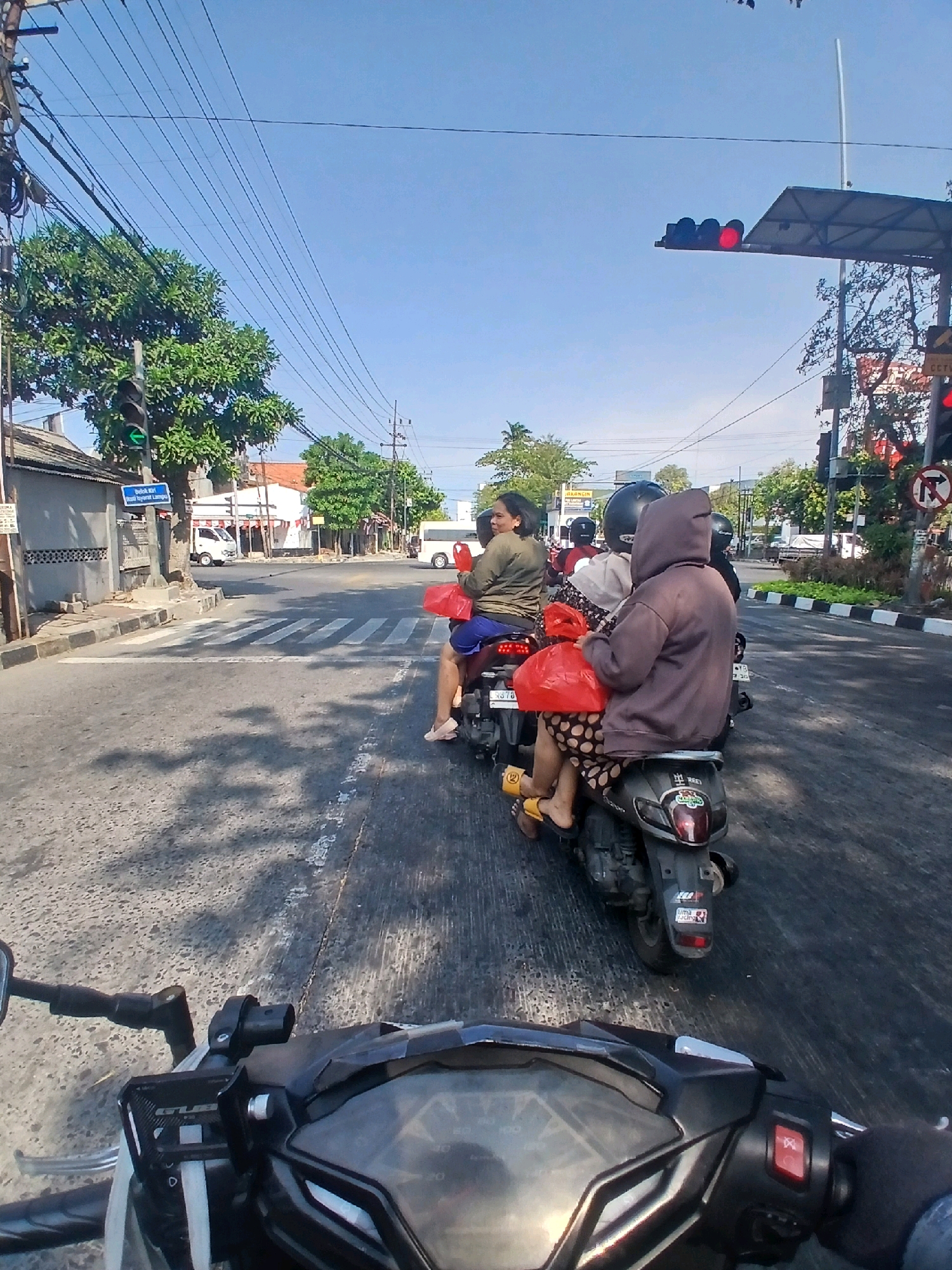 This image captures a scene at an intersection in Surabaya, Indonesia, on a clear day. The perspective is from a motorcycle rider, looking forward. Three motorcycles are visible, each carrying two passengers.
On the closest motorcycle to the viewer, the rear passenger is a person wearing a brown hooded sweatshirt and a patterned skirt, holding a red plastic bag. The person in front of them is wearing a green top and blue shorts, also holding a red plastic bag. The second motorcycle from the viewer has two passengers, one wearing a black helmet. The third motorcycle also has two passengers, with the rear passenger wearing a helmet.
The intersection appears to be controlled by traffic lights. A traffic light with a green arrow is visible on the left, and a traffic light with red lights is visible overhead on the right. A pedestrian crossing is marked on the road. The environment includes buildings on the left side of the street, trees, and overhead power lines. The road surface is asphalt with some visible markings and shadows.