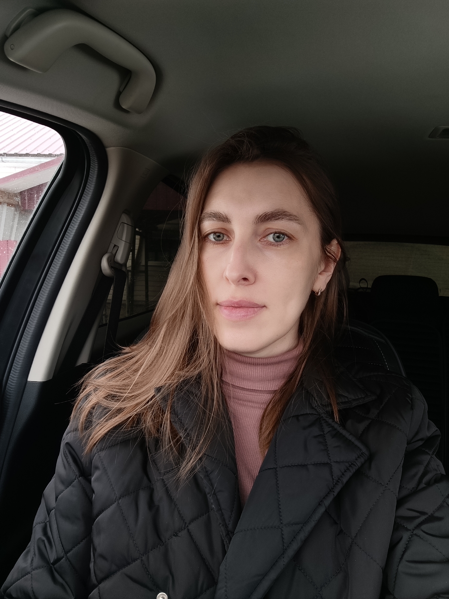 A woman with long brown hair and light eyes sits in a car. She is wearing a pink turtleneck sweater and a black quilted jacket. The car interior is black and grey. Outside the car window, a building with a red roof is visible, suggesting a possible residential or commercial area. The location is Фоки, Russia.