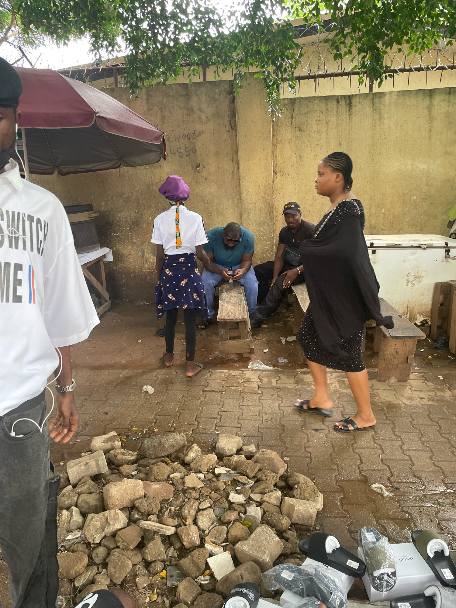 The image depicts an outdoor scene in an urban environment that appears to be in Abuja, Nigeria. In the foreground, a person wearing a white t-shirt with "SWITCH ME" printed on it and dark pants is partially visible, facing away from the camera. To their left, a pile of rough, angular rocks and debris lies on a paved ground.
Behind the foreground person, several other individuals are present. A woman in a black dress adorned with rhinestones is walking towards the right side of the frame. Further back and to the left, a person wearing a purple headwrap and a skirt stands facing away from the camera. A man in a blue t-shirt and jeans is seated on a wooden bench, holding a mobile phone. Another man, wearing a black t-shirt and a black cap, is seated beside him, also looking down.
An umbrella with a dark red and grey canopy is open to the left of the frame, casting a shadow. The background features a textured, yellowish wall with some graffiti or markings. To the right, a large, white refrigeration unit is visible. The ground is a mix of paved interlocking stones and dirt, with some areas appearing wet. Green foliage from trees overhangs the scene from above. The overall impression is a casual gathering or marketplace setting in an outdoor, somewhat unkempt urban area.
