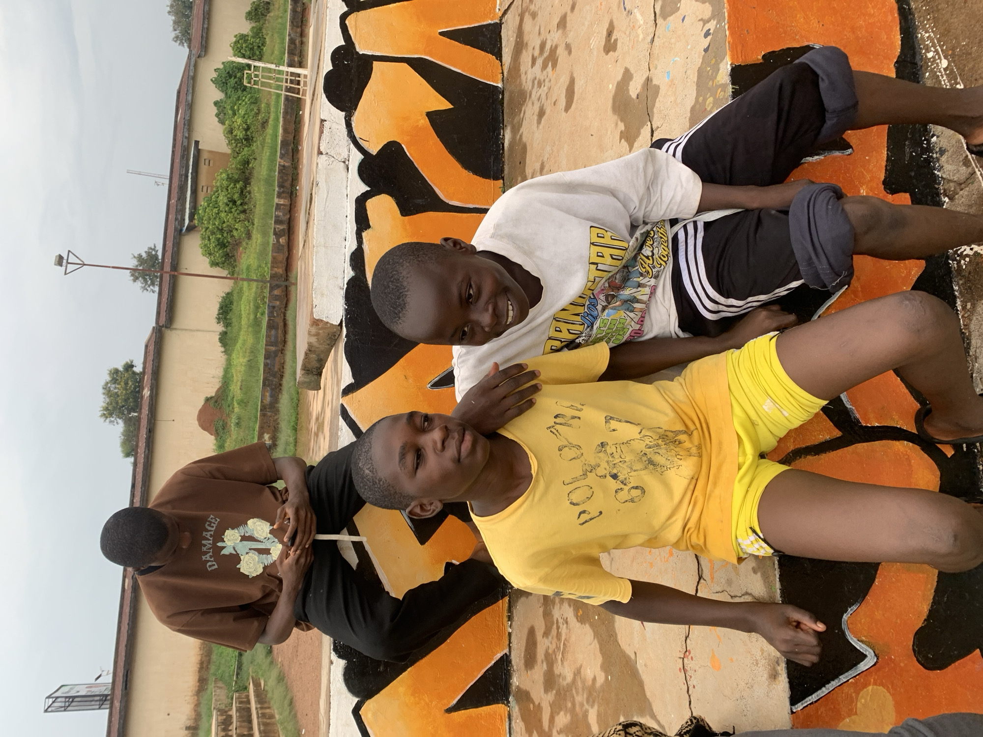 In Gura Top, Nigeria, three young Black males are seated outdoors on a tiered, graffiti-covered concrete structure under an overcast sky. In the background, a tan building with a reddish-brown roof is visible, along with overgrown green vegetation and a single lamppost. The male in the back, wearing a brown t-shirt and black pants, is looking down at his phone. Two other males are seated in the front. The one on the left, wearing a yellow t-shirt and yellow shorts, has his arm around the shoulders of the male to his right. The male on the right, wearing a white t-shirt with graphics and black pants with white stripes, is smiling and looking to his left. The structure they are sitting on is adorned with orange and black graffiti.