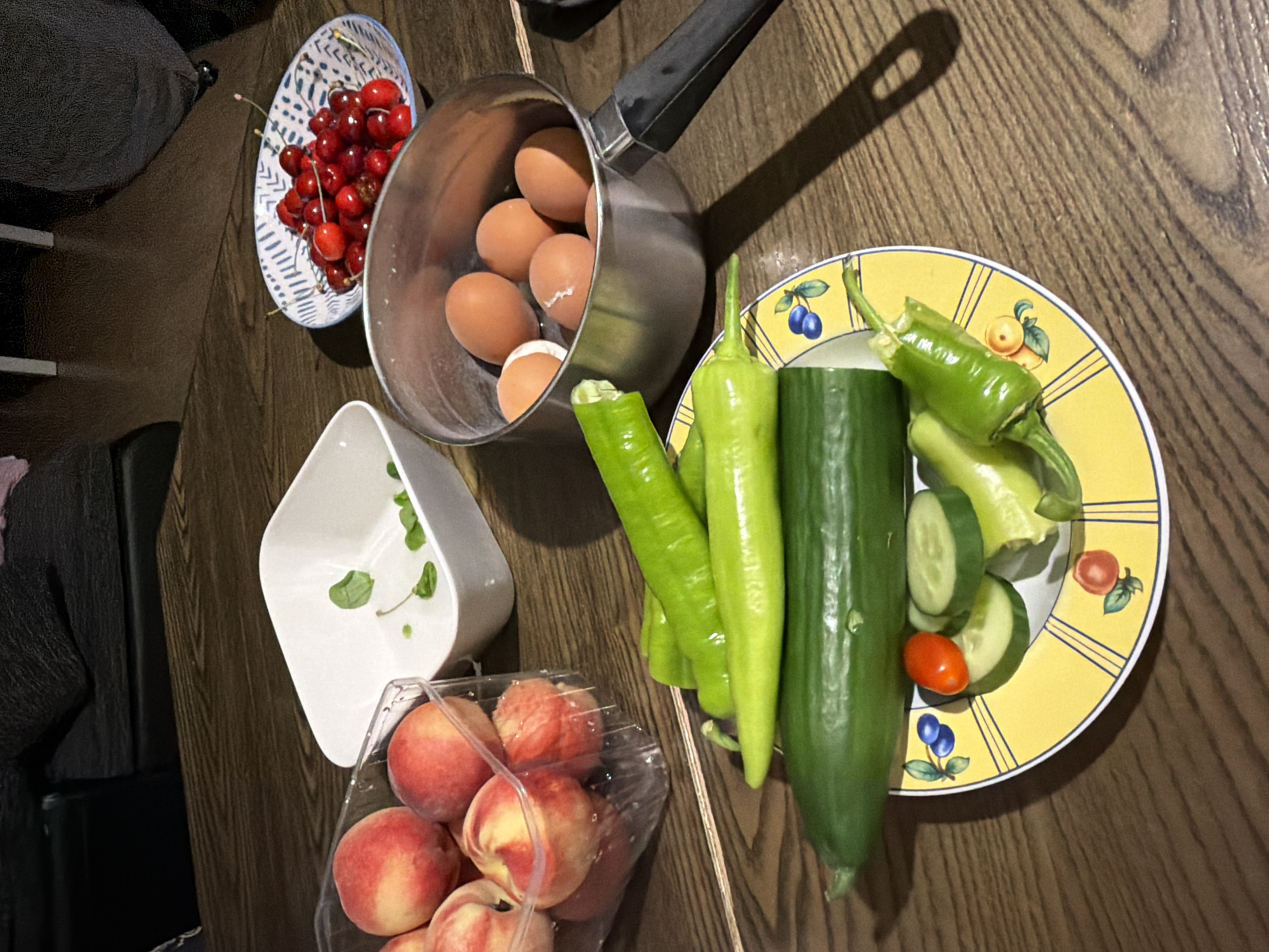 An overhead shot displays a variety of fresh food items and cooking ingredients arranged on a warm-toned wooden table. The scene suggests preparations for a meal or a collection of groceries.
In the upper left, a white ceramic bowl adorned with blue geometric patterns holds a generous cluster of bright red cherries, complete with their green stems. Adjacent to this, a silver saucepan contains several brown eggs; at least six are visible, and some appear to have slight cracks, possibly from being boiled or prepared for cooking.
Towards the bottom left, a clear plastic container is filled with round, ripe peaches, exhibiting a soft blend of yellow and reddish hues. Above the peaches, a small, white, irregularly shaped bowl holds a few green mint leaves.
Dominating the right side of the image are fresh green vegetables. A long, dark green cucumber lies horizontally, and several vibrant green peppers, some long and slender, others shorter and broader, are scattered around. A round yellow plate, decorated with illustrations of blue and orange fruits, holds a medley of sliced cucumber, a small red cherry tomato, and pieces of green pepper, indicating some of the vegetables have already been prepared.
The overall setting is indoors, likely a kitchen or dining area, with the wooden surface serving as a backdrop. There are no individuals visible, no explicit time of day, weather, or location cues within the image itself to suggest Strasbourg, France, beyond the given context. No visible text is present.