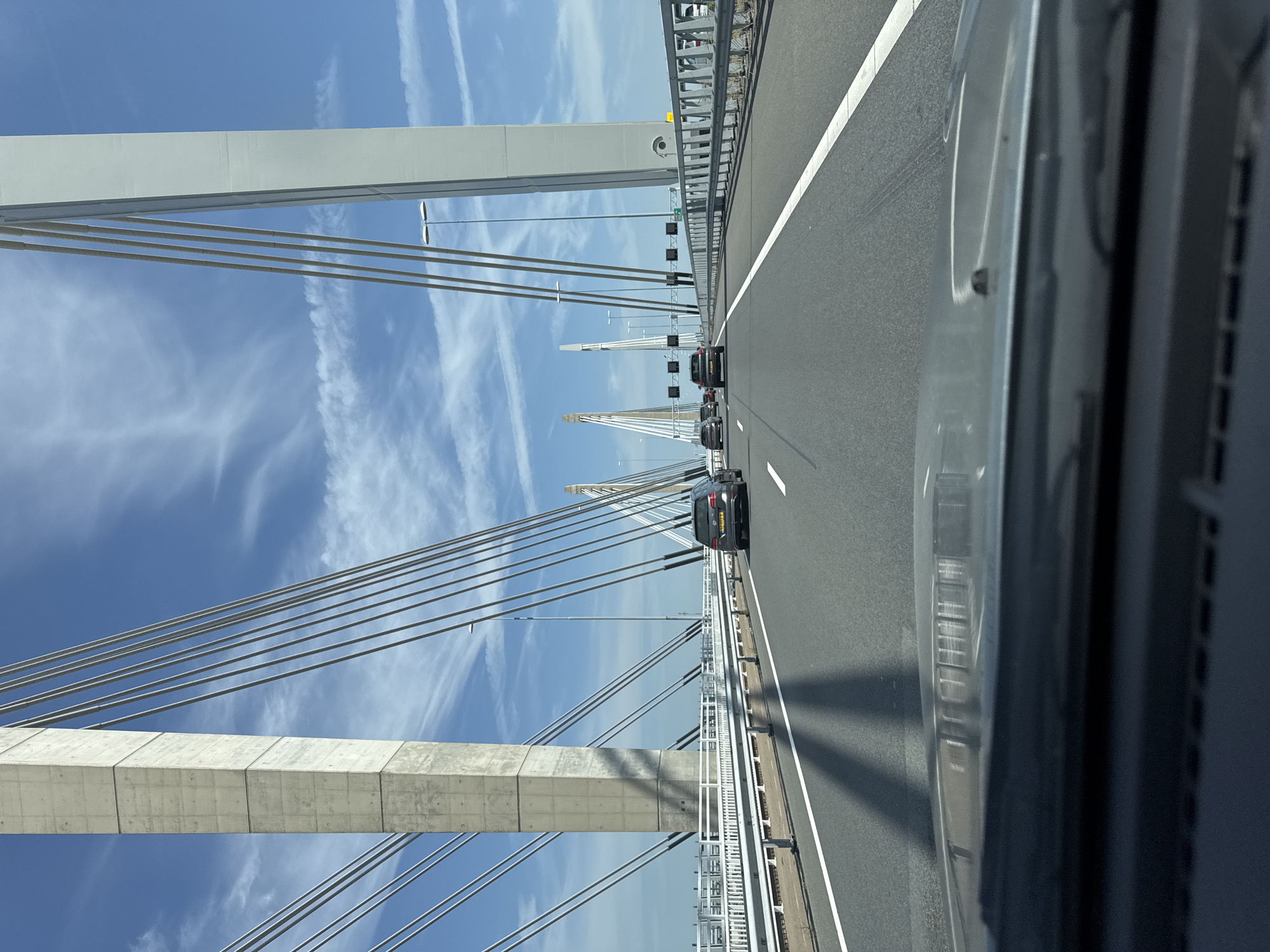 The image captures a view from a vehicle driving on a multi-lane cable-stayed bridge, likely in the vicinity of Andelst, Netherlands, given the distinctive architecture of the bridge towers. The sky above is a clear blue with scattered white clouds. The bridge features prominent concrete and white pylons from which numerous cables extend to support the roadway. Several cars are visible on the road, all traveling in the same direction as the viewer's vehicle. The road surface is asphalt with white lane markings. The foreground shows the dashboard and windshield of the vehicle, with reflections of the bridge structure and sky visible on the windshield. The environment is characterized by the vastness of the bridge structure against the open sky, suggesting a location over water or a significant geographical feature.