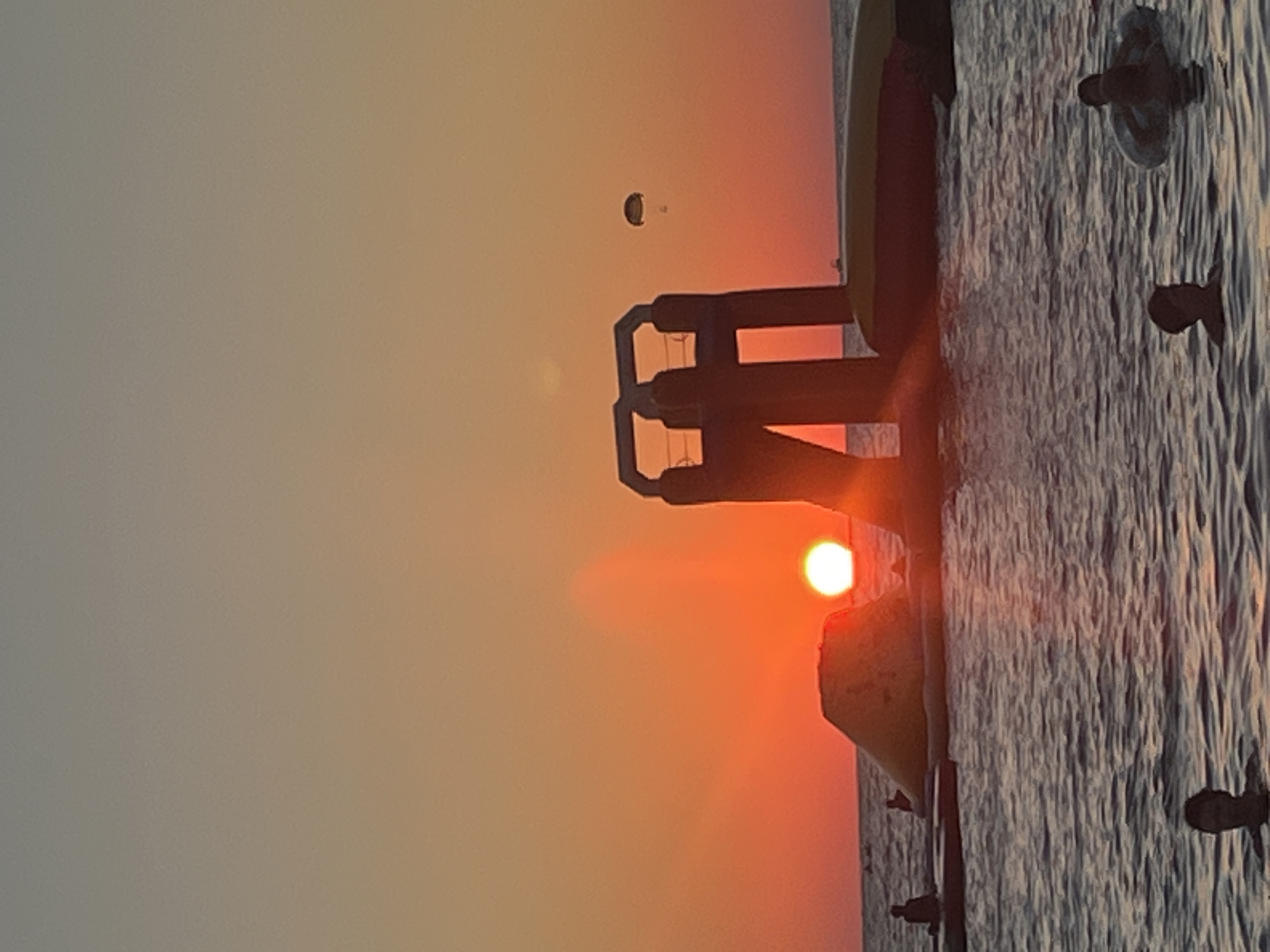 The image depicts a serene sunset over the Black Sea in Sochi, Russia. The sky is a gradient of warm oranges and yellows, with the sun a bright, intense orb just above the horizon. In the foreground, the calm, rippling water reflects the sunset's hues. Several people are visible in the water, some swimming and others relaxing on inflatable rings.
Further out in the water, there is an inflatable water park structure, featuring slides and platforms, illuminated by the fading sunlight. A small boat can be seen in the distance near the horizon. In the sky, a paraglider is visible, adding a touch of recreational activity to the scene. The overall atmosphere is tranquil and picturesque, capturing a typical summer evening on the coast of Sochi.