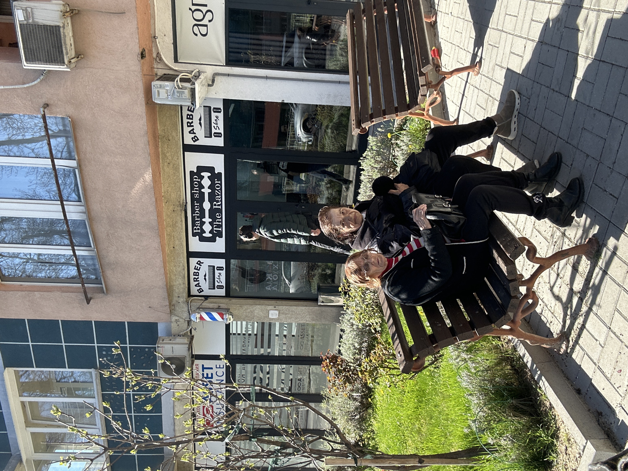 Here's a detailed description of the image:
**General Description:**
The image shows two people sitting on a bench on a city street. They are located in front of shops and offices in Sofia, Bulgaria.
**People:**
* Two people are on the bench.
* One is an older woman, seen smiling and looking towards the camera. She is wearing a dark jacket and appears to have short blonde hair.
* The second person is partially obscured, but appears to be wearing dark clothing as well.
* There are reflections of other people in the shop windows.
**Objects:**
* A brown wooden bench with decorative metal legs.
* A black handbag is on the bench next to the woman.
* Visible storefronts with signs, including a barber shop named "The Razor", and an insurance agency. A barber pole is also visible.
* Air conditioning units are attached to the building facade.
* Windows with metal bars are present.
* Green grass and low shrubbery are near the bench.
**Environment:**
* The location is a city street in Sofia, Bulgaria (based on storefront signage in Cyrillic script).
* The street is paved with stone.
* A tree with sparse new growth is in the foreground.
* The day appears sunny, with shadows cast on the ground.
* The architecture suggests an urban area with commercial buildings.
**Actions/Interactions:**
* The two people are sitting on the bench.
* One person is looking at the camera, possibly posing for a photo.
* People are reflected in the shop windows, suggesting foot traffic on the street.