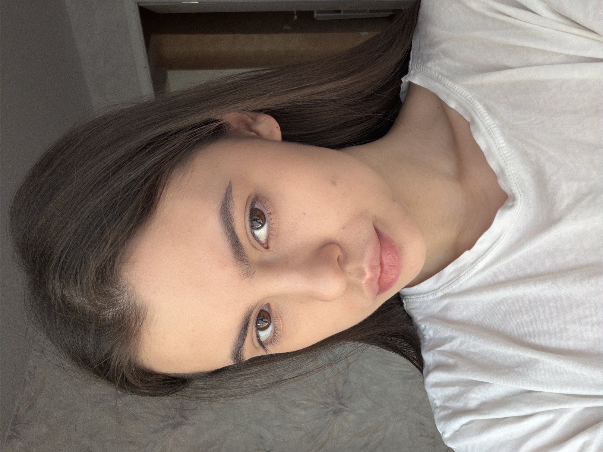 The media file displays a close-up portrait of a young woman, her head positioned on the left side of the frame and her body extending to the right, suggesting a reposed or tilted posture. She has long, straight dark brown hair that cascades around her face and extends across the frame. Her face is angled slightly towards the viewer, with her brown eyes looking directly into the camera. She has distinct, well-groomed dark eyebrows and full, naturally pink lips. Her complexion appears clear, with a few small moles visible on her cheek and near her ear. She is wearing a simple white crew-neck t-shirt, visible on the right side of the image with soft folds in the fabric.
The background is softly blurred and indistinct, indicating an indoor setting. A darker, possibly textured surface is visible beneath her head, and a darker, architectural element like a wall or corner appears in the upper left background. The lighting is soft and even, highlighting her features without harsh shadows, suggesting natural or diffused indoor light. Her expression is neutral and calm, conveying a sense of quiet observation. There is no visible text in the image. The photo captures a moment of gentle contemplation. The location context provided is Chaikovsky, Russia, although no specific visual cues within the image confirm this.