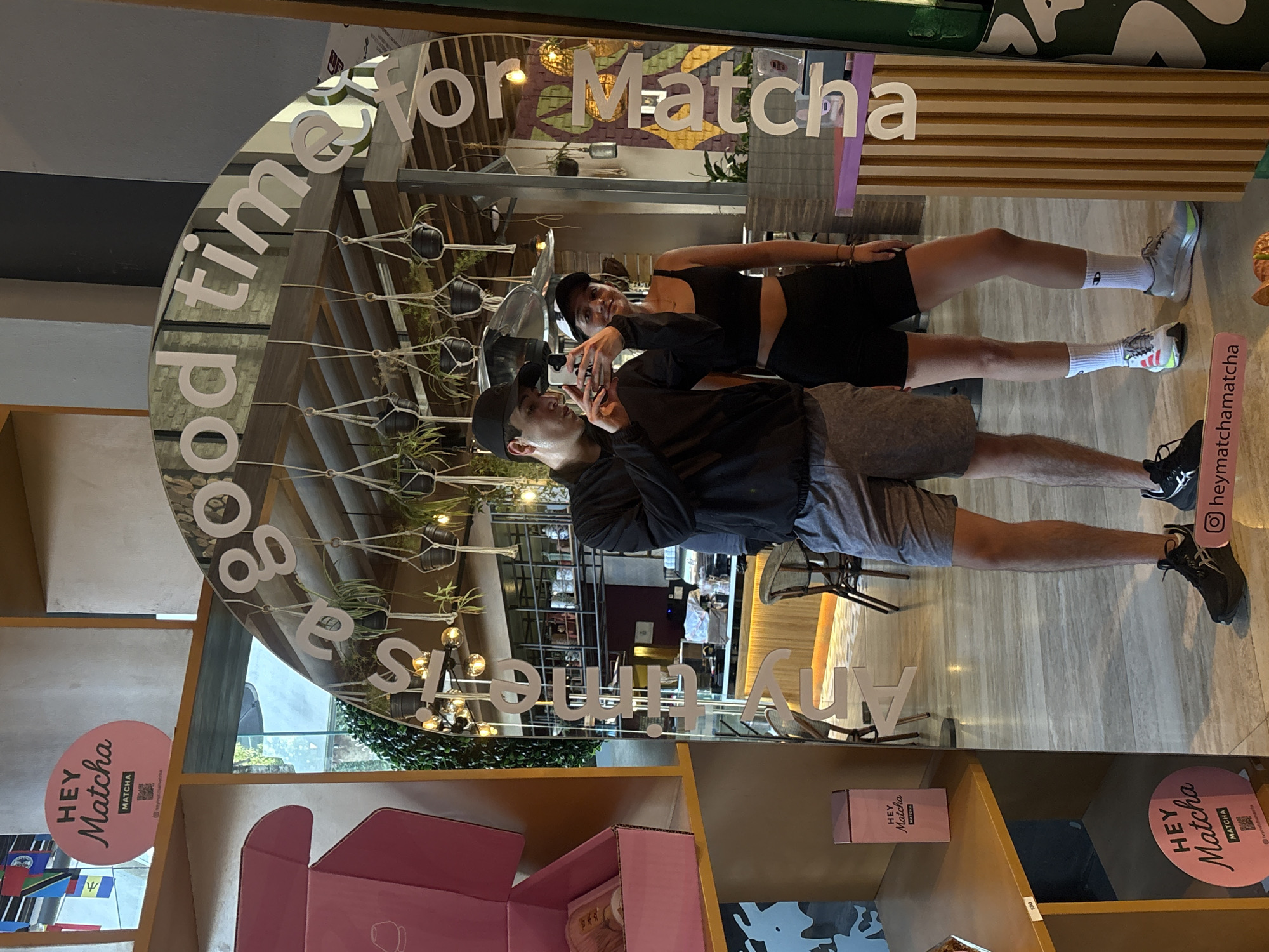 Here is a forensic description of the image:
**Scene:**
The image captures a scene at "Hey Matcha," a business located in San Bernardino Tlaxcalancingo, Mexico. A large, curved mirror is the focal point, displaying the phrase "Any time is a good time for Matcha." The mirror's placement allows reflections of the interior of the establishment.
**People:**
Two individuals are visible, presumably customers, taking a photo with the mirror. One person is in the foreground, holding a smartphone to take the picture. They are wearing a dark cap, a dark jacket, and grey shorts. The second person is partially visible behind them, dressed in a black top and black shorts.
**Objects:**
Several "Hey Matcha"-branded items are present, including boxes and signage. Various decorative elements, such as plants and lighting fixtures, are integrated into the design.
**Environment:**
The environment appears to be a modern, well-lit commercial space. The interior design includes wooden elements and a neutral color palette. The reflections in the mirror suggest an open floor plan with seating areas.
**Action:**
The action observed is the individuals taking a photo, likely for social media or personal documentation of their visit to "Hey Matcha."