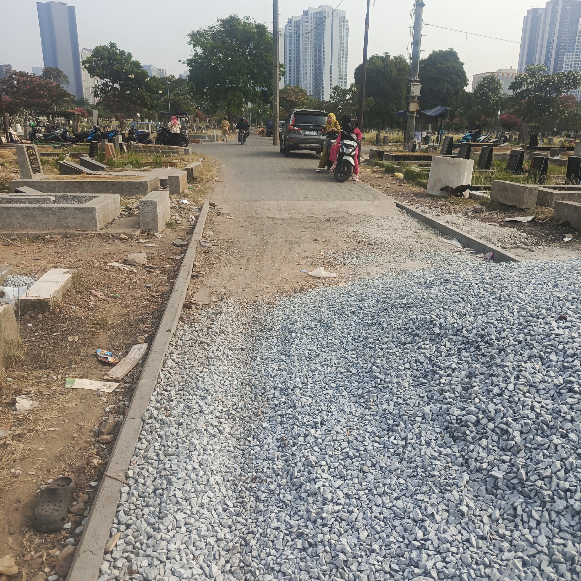 The image depicts an outdoor scene at a cemetery in South Jakarta, Indonesia. In the foreground, a large pile of grey gravel is visible, occupying the right half of the frame and extending towards the bottom right corner. A concrete curb separates this gravel from a dusty, unpaved path. On the left side of the path, there are several concrete grave structures, some partially filled with gravel, and remnants of building materials. A dark-colored rubber clog is discarded on the ground to the left.
Further into the scene, a paved road runs through the cemetery, lined with more grave sites and vegetation. Several motorcycles are parked along the left side of the road, near some of the graves. Two women, one wearing a yellow hijab and the other a pink hijab, are seated on a pink and white motorcycle on the right side of the road. Behind them, a grey SUV with a license plate starting with "B" is stopped on the road. Two other individuals are visible on motorcycles further down the road. In the background, tall apartment buildings and trees are present, indicating an urban setting. The sky is hazy and overcast.