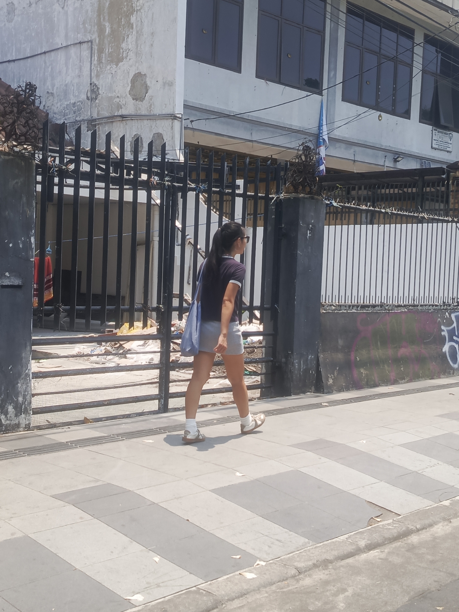 The image depicts a woman walking on a sidewalk in Surabaya, Indonesia. She is wearing a dark t-shirt with white trim, grey shorts, white socks, and light-colored sandals. A light blue bag is slung over her left shoulder. The background shows a black metal fence with barbed wire on top, and behind it, a dilapidated building with peeling paint. Rubble and debris are visible near the fence. The sidewalk is paved with alternating light and dark gray square tiles, and a tactile paving strip runs along its edge. The overall environment appears to be an urban setting, possibly an industrial or less well-maintained area of the city.