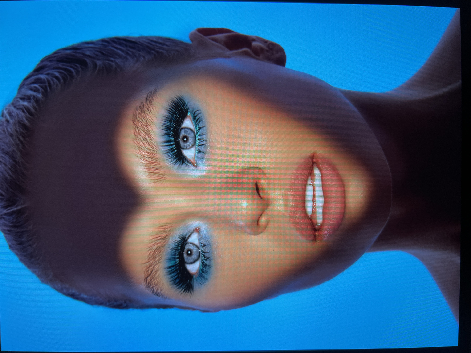 The image is a close-up, head-and-shoulders portrait of a woman with striking makeup. She has on vibrant teal eyeshadow and dramatic false eyelashes. Her lips are a peachy pink, slightly parted to reveal her teeth. A heart-shaped shadow falls across her forehead and eyes, obscuring part of her face. The background is a solid, bright blue. The image's composition and lighting suggest a studio setting for a fashion or beauty photoshoot. There is no information in the image to suggest a location in Saint Petersburg, Russia.
