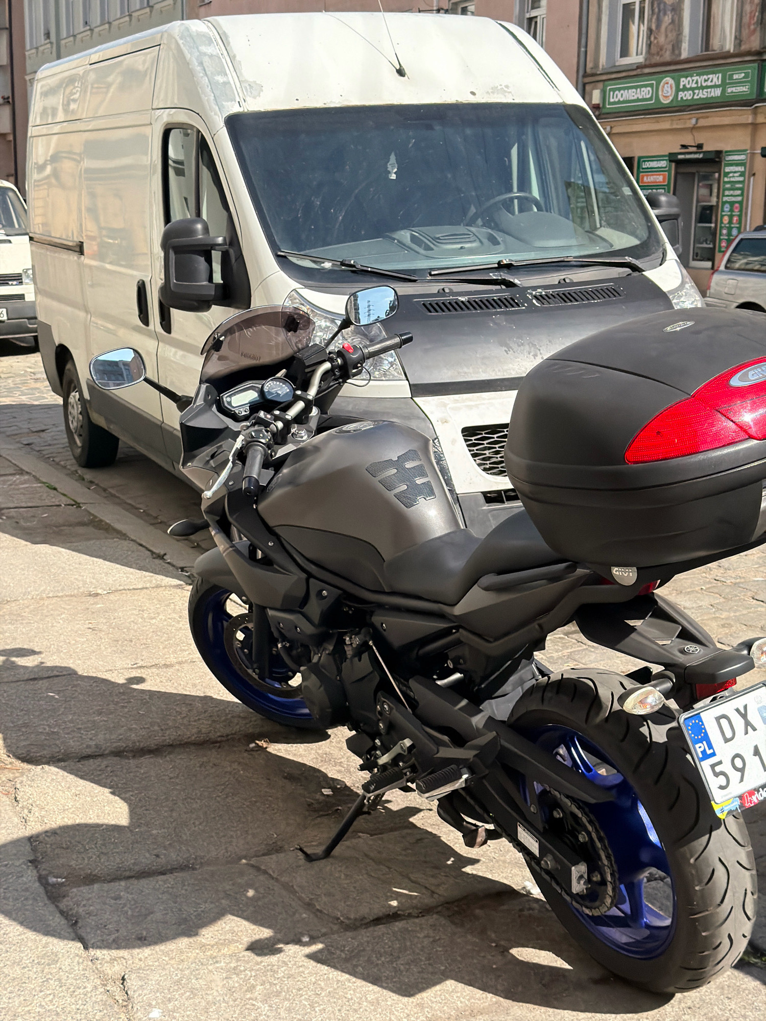 A dark gray Yamaha motorcycle is parked on a cobblestone street in front of a white van. The motorcycle has a black Givi top case, blue rims, and a license plate from Poland that reads "DX 591". In the background, a building with signs for "LOOMBARD" and "POŻYCZKI POD ZASTAW" is visible, indicating a pawn shop and loan service. The scene is illuminated by sunlight, casting shadows on the ground. The environment suggests an urban setting in Wrocław, Poland. There are no people visible in the image.