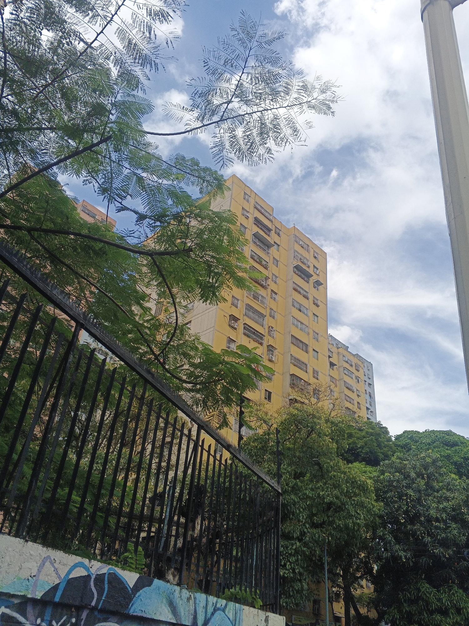 The image captures a scene in Caracas, Venezuela, featuring a yellow apartment building viewed from street level. A wrought iron fence and a graffiti-covered wall are in the foreground. Trees with green foliage partially obscure the view of the building and the sky, which is streaked with white clouds. A portion of a concrete pillar is visible on the right side of the frame. No people are visible in the image.