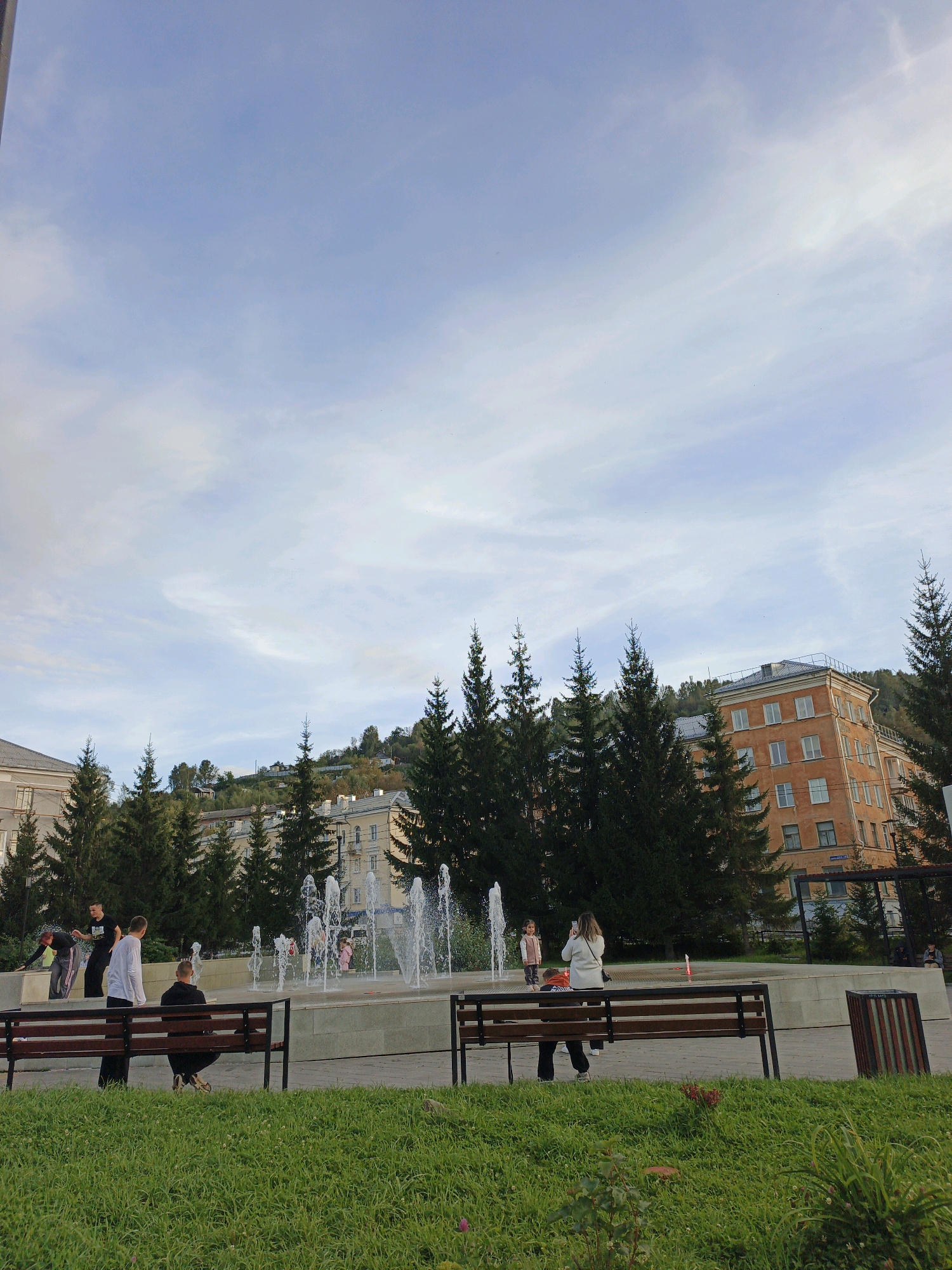 The image depicts a public park area in Zlatoust, Russia, featuring a fountain with multiple jets of water spraying upwards. Several individuals are present in the park. In the foreground, two park benches are visible, with one occupied by a person sitting and facing away from the camera, and another with a person sitting and facing the fountain, their back to the camera. Further back, near the fountain, a group of younger people are interacting. One person is standing and appears to be taking a photograph with a mobile device, while another individual, a child, stands near the fountain. Behind the fountain, a line of tall evergreen trees forms a backdrop. Beyond the trees, residential buildings with orange and light-colored facades are visible, situated on a gentle slope. The sky above is a light blue with scattered white clouds. The overall scene suggests a pleasant outdoor environment in a city setting.