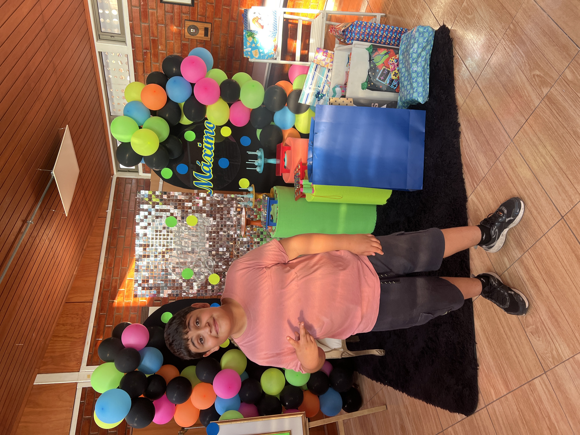 A boy is lying on a black rug in front of birthday decorations. The decorations include a balloon arch of black, pink, blue, green, and orange balloons, a sign reading "Maximo" in yellow letters, and a metallic tinsel backdrop. Behind the boy are gifts in bags and boxes. The setting appears to be indoors, with a brick wall and wood-paneled walls visible. The floor is tiled. The location is Arica, Chile.