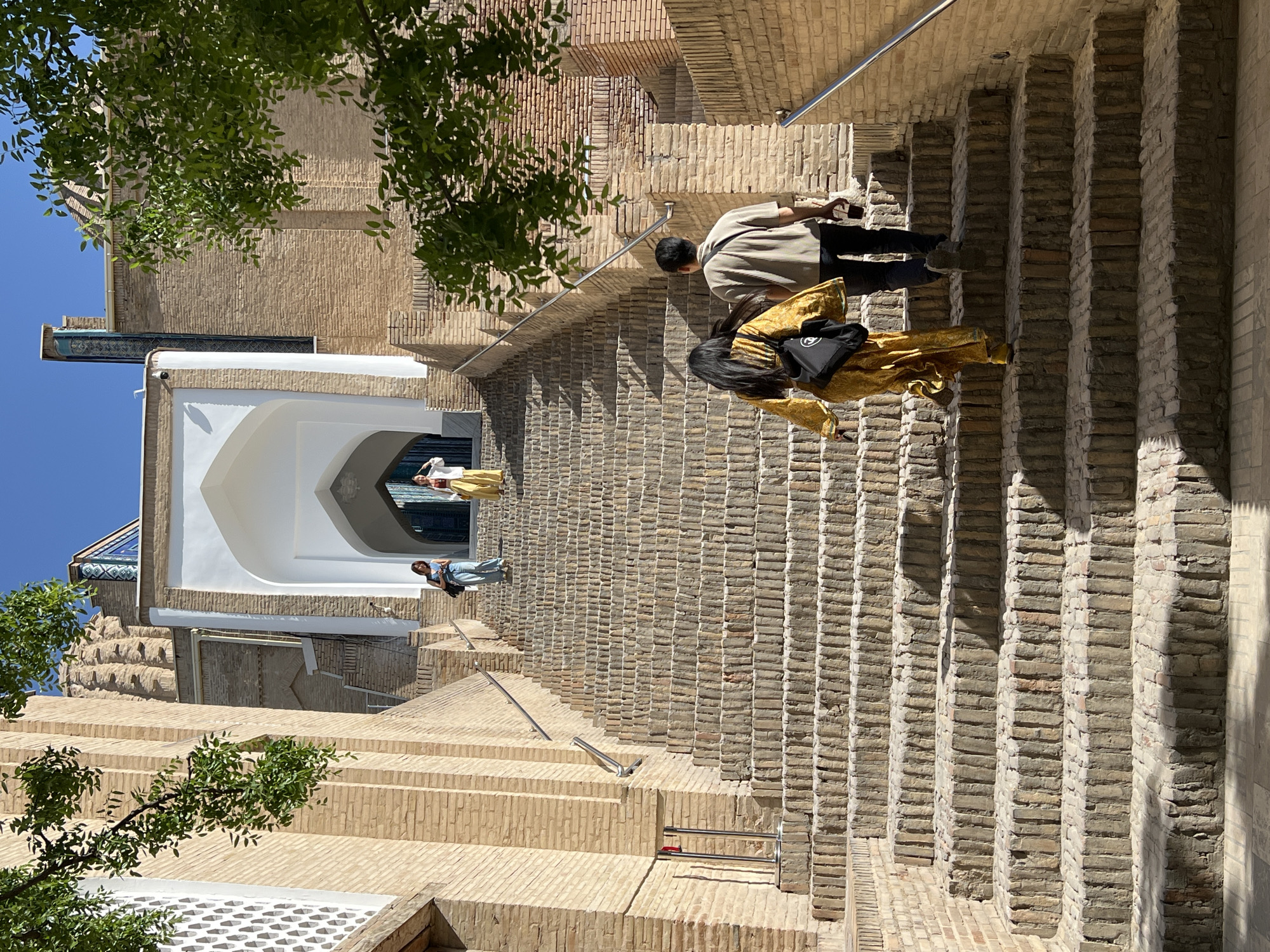 The image shows a wide, ancient stone staircase leading up to an arched entrance of a historic building. The stairs are made of rough-hewn stone blocks, weathered and worn, with a textured surface. The walls flanking the staircase are constructed from aged, light-brown bricks, showing signs of age and history.
Several people are visible on the stairs. Two individuals are ascending the stairs in the foreground, a woman in a flowing yellow garment with a black bag slung over her shoulder, and a man in a light-colored shirt and dark trousers. Further up the stairs, two more individuals are also ascending, one in a blue top and jeans, and another in a white jacket and yellow trousers. Above them, at the arched entrance, another person can be seen.
The architecture suggests a location with a rich historical and cultural heritage. The overall atmosphere is one of exploration and discovery, set against a backdrop of timeless structures. The scene is lit by bright sunlight, casting shadows on the stairs and highlighting the textures of the stone and brick. This setting is consistent with the architectural style and ambiance found in historical sites within Samarkand City, Uzbekistan, such as the Registan complex or the Shah-i-Zinda necropolis, known for their intricate brickwork and grand staircases.