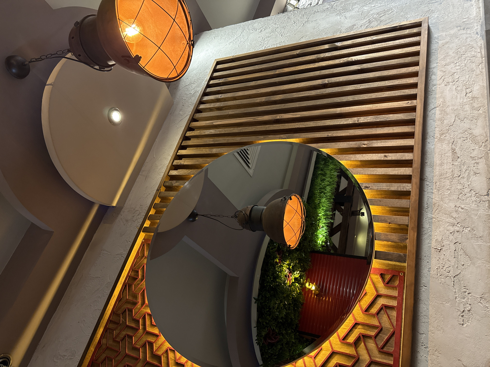 The image captures an interior scene, likely a commercial establishment such as a restaurant or cafe, located in Unknown, Unknown. A prominent industrial-style pendant lamp, made of dark, aged metal with a protective wire cage, hangs from the ceiling in the upper right quadrant, emitting a warm orange glow from its exposed bulb. The ceiling is multi-leveled, featuring a dark grey section and a lighter, arched recess containing a small, bright white recessed spotlight. On the left side, a large circular mirror with a decorative frame is mounted on a wall. The frame displays an intricate, geometric pattern in yellow and red. The mirror reflects an internal area featuring a similar pendant lamp, a wall adorned with green artificial foliage, a red slatted structure (possibly a shutter or blind), and an overhead air vent. Adjacent to the mirror, on the right, is a rectangular wall panel composed of numerous vertical wooden slats of varying shades. The wall surfaces visible are textured, appearing as rough plaster or concrete, in a light, neutral gray tone.