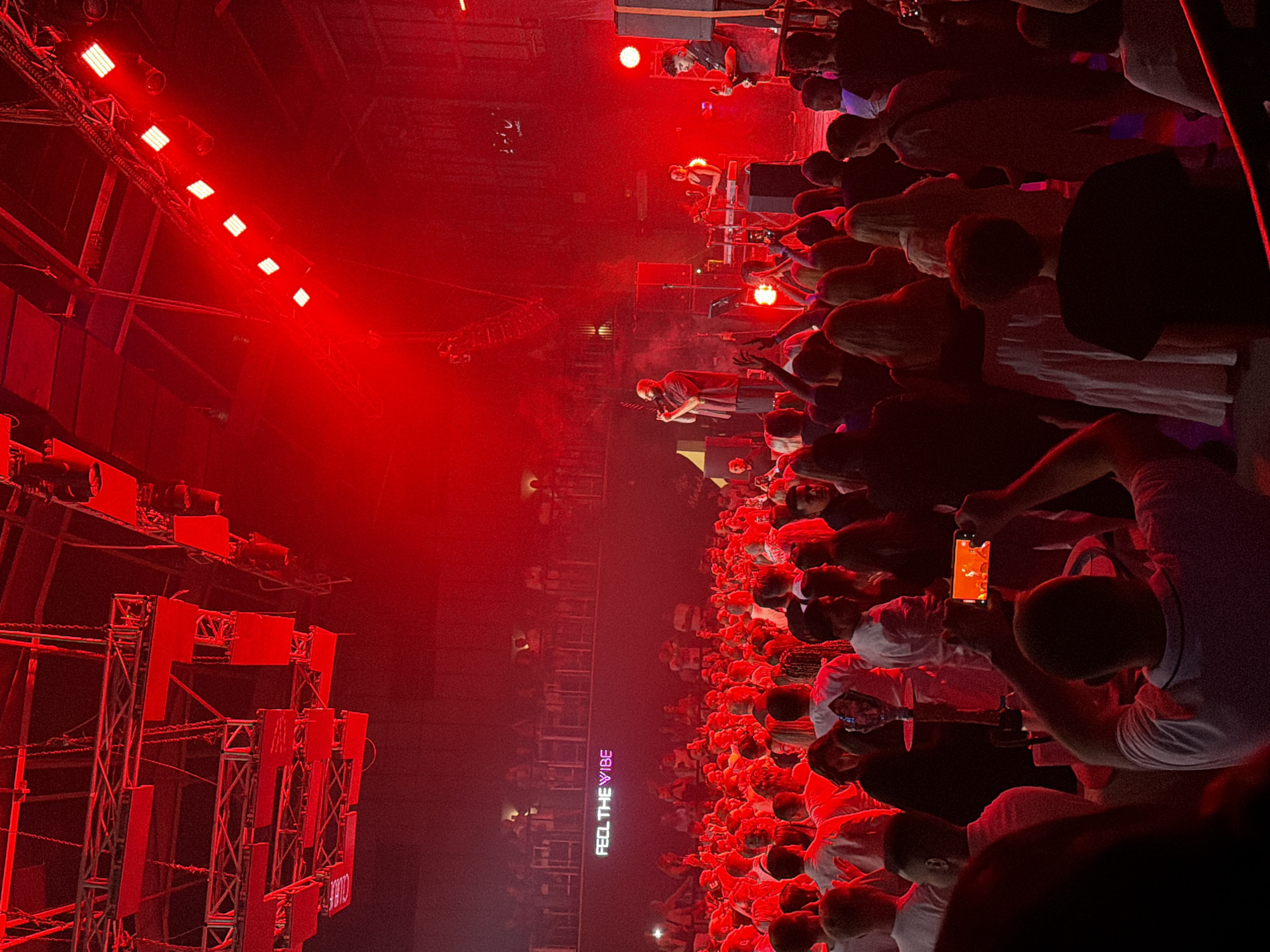 The image depicts a lively indoor concert or club scene, bathed in intense red lighting, in Sharm El Sheikh, Egypt. A crowd of people is gathered, many facing a stage where performers are active. On stage, a man in a red and black patterned shirt stands at a microphone, appearing to sing or speak, while another individual plays a keyboard and a third plays a guitar. Behind the performers, equipment and speakers are visible. Above the crowd, a sign illuminated with purple and white lights reads "FEEL THE VIBE." The venue appears to have multiple levels, with balconies or upper seating areas visible in the background. The overall atmosphere is energetic and engaging, with concert-goers actively participating in the event, some holding up phones to record.