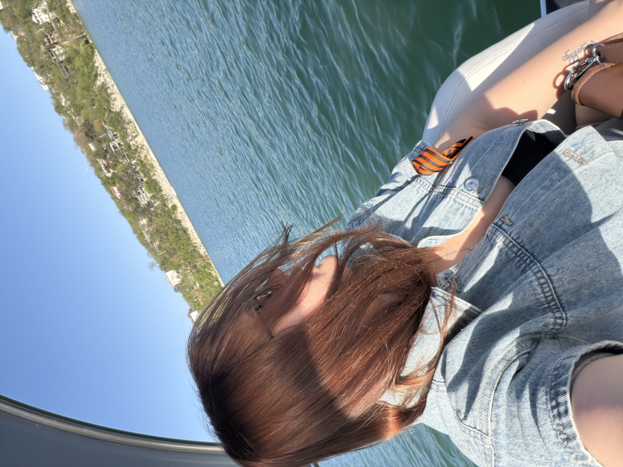 A woman with long, auburn hair is on a boat on Abrau Lake in Abrau-Dyurso, Russia. She is wearing a denim jacket over a black top, with a St. George ribbon pinned to the jacket. A handbag is near her arm, which has a tattoo on it. The lake water is blue-green, and the opposite shore is visible in the background. The sky is clear and blue.