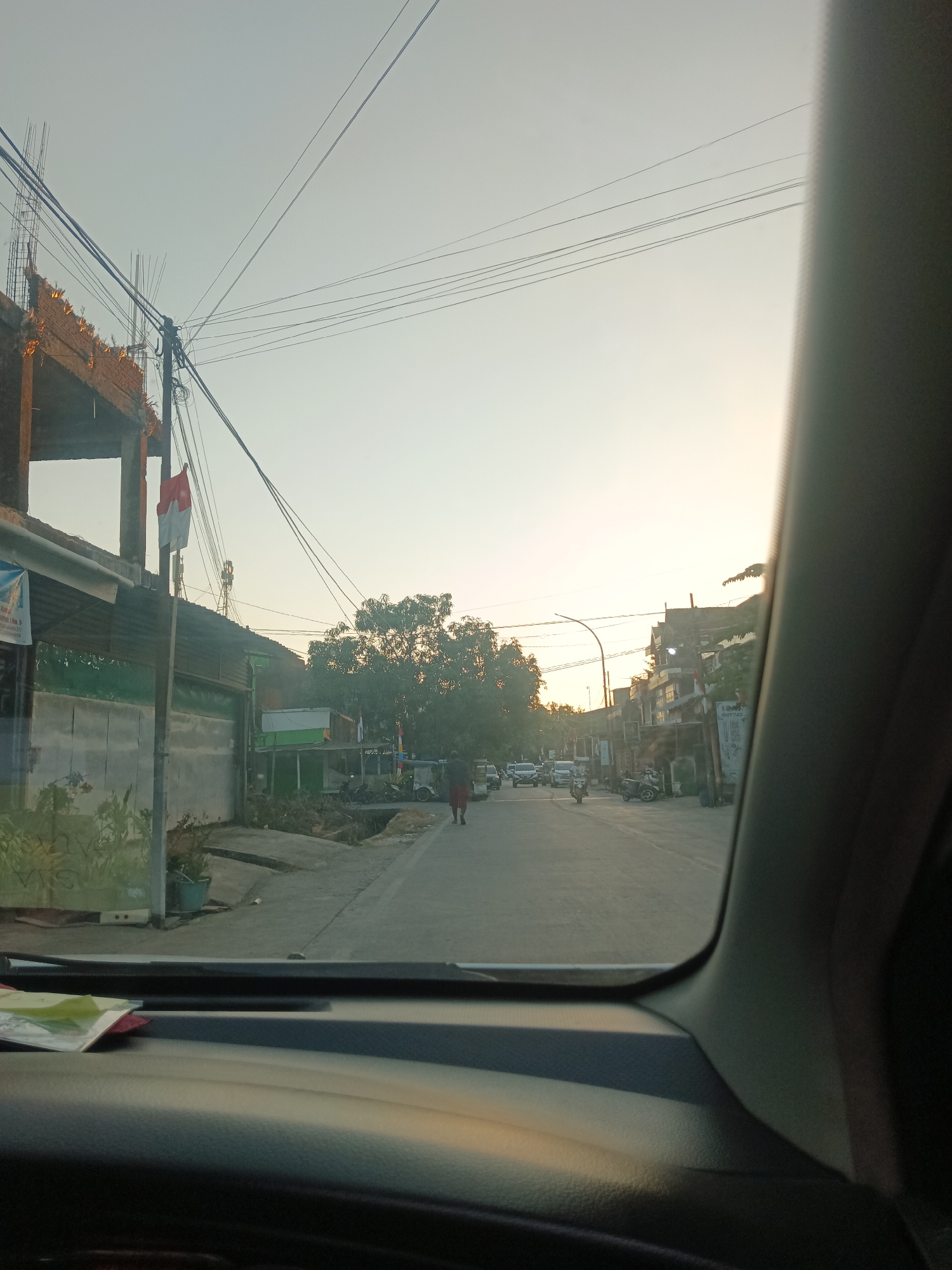 The image captures a view from inside a vehicle in Pangkajene, Indonesia, looking out at a street scene during what appears to be late afternoon or early morning. The sky is bright with a pale yellow hue, suggesting sunrise or sunset. On the left side of the road, a building under construction is visible, with exposed rebar at the top. A flag, appearing to be the Indonesian flag with its red and white stripes, is planted near this building. Adjacent to the construction site are several single-story structures with corrugated metal roofs and signs. The street itself is paved and shows moderate traffic, including cars, motorcycles, and a pedestrian walking away from the camera. Trees are present along the roadside, and power lines crisscross the sky. The foreground of the image is dominated by the interior of the vehicle, with a dark dashboard and part of the car's pillar.