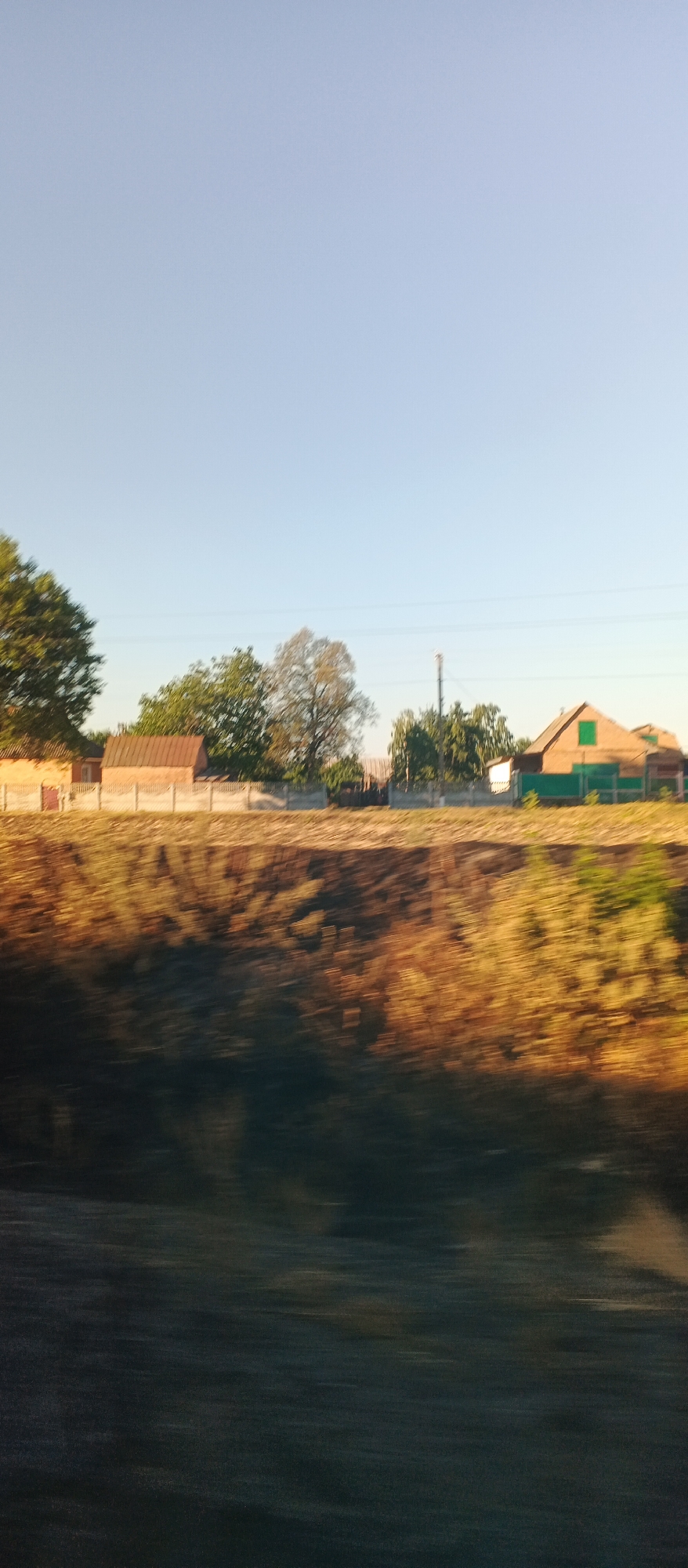 This is a motion-blurred image of a rural scene captured in Khlystunivka, Ukraine. The sky is clear and pale blue. A line of trees and houses is visible in the midground, separated from the foreground by a low concrete fence. The houses appear to be simple, rural structures, some with visible roofs. The foreground is dominated by a dark, out-of-focus area, possibly a body of water or a shadowed area of dense vegetation. The overall impression is of a moving vehicle's perspective, looking out at the landscape.