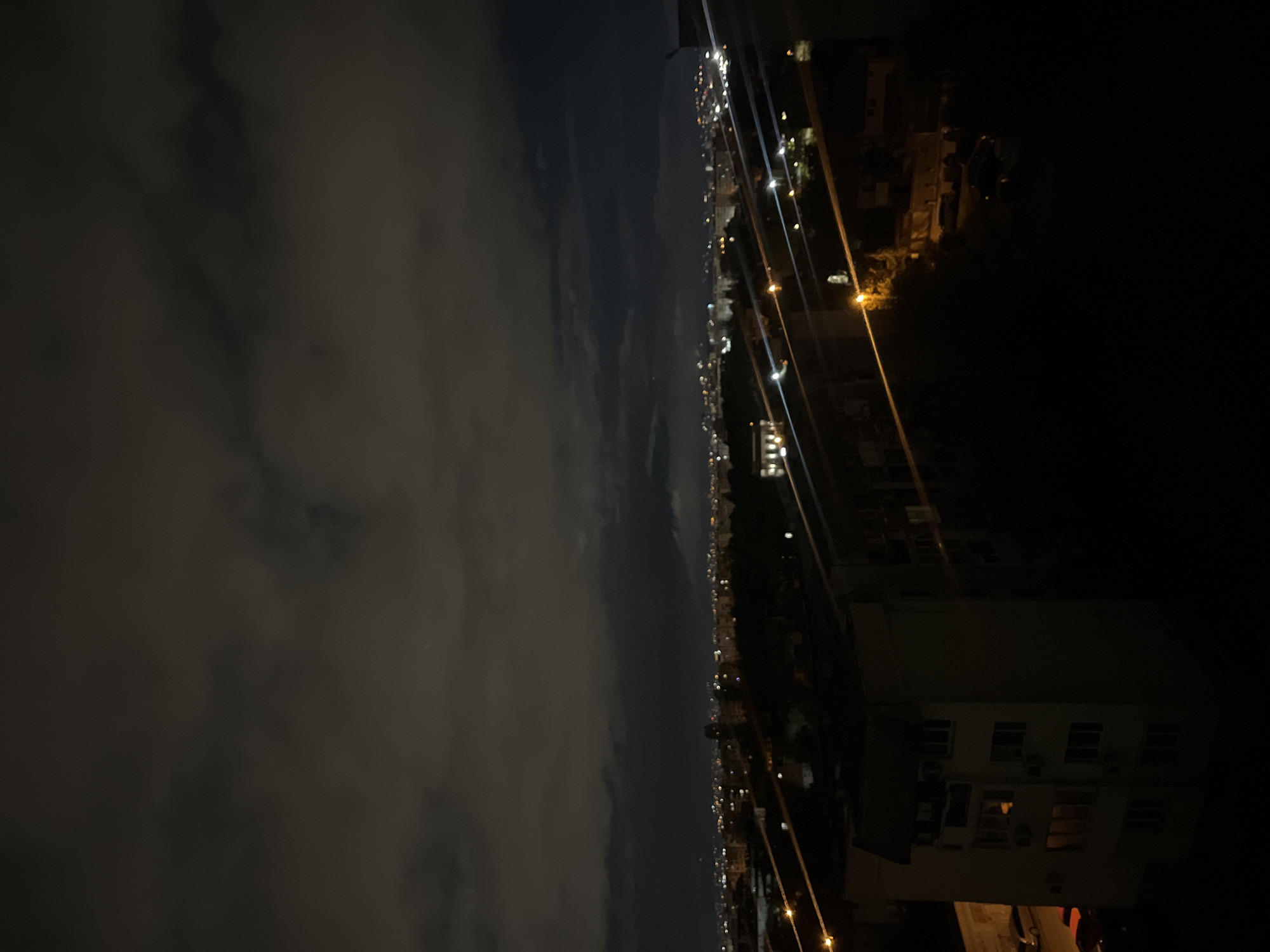 The image shows a nighttime view from a high vantage point, possibly a building in Sofia, Bulgaria. The left side of the image is dominated by a dark, cloudy sky. A line of city lights stretches horizontally across the middle of the image, indicating a distant cityscape. On the right side, a building is visible in the foreground, with illuminated windows. Some power lines are crossing through the view.
