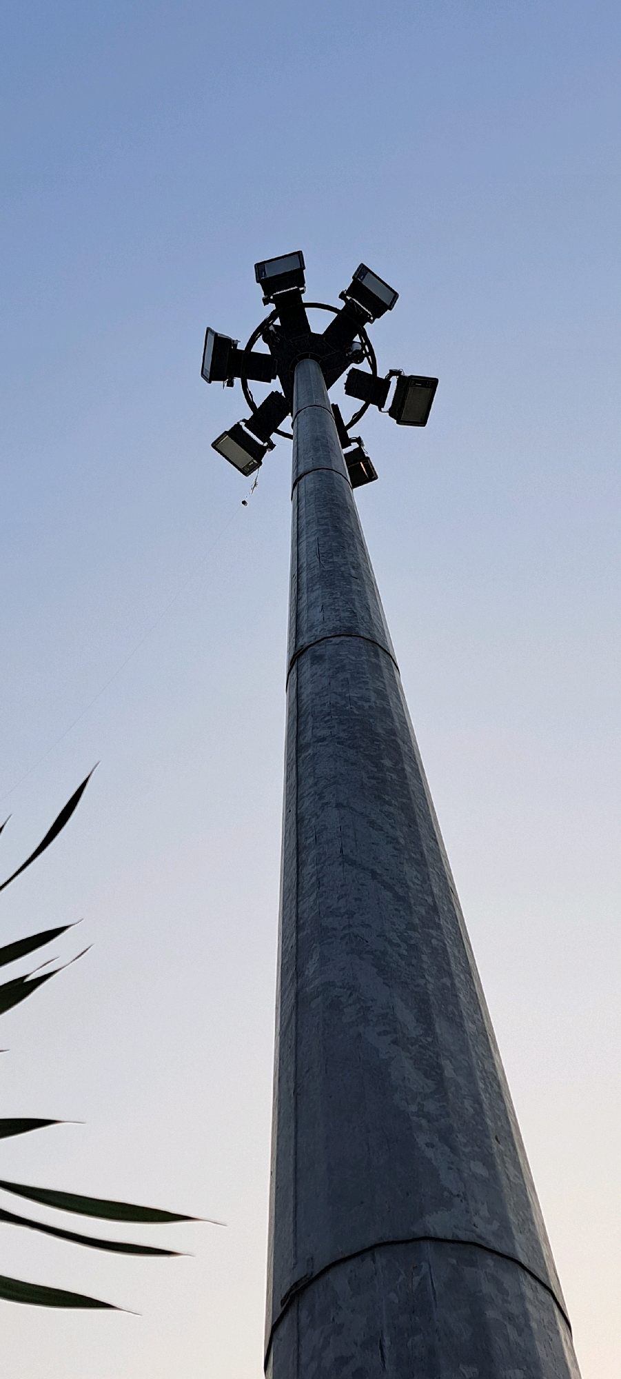 The image shows a tall, grey, metallic lamppost with multiple light fixtures at its top, set against a clear, pale blue sky. The lamppost is cylindrical and appears to be made of segmented metal panels. It tapers slightly towards the top. There are several rectangular light fixtures arranged in a circular pattern around the top of the pole. Some of these lights are directed downwards, while others are angled differently. In the lower left of the frame, the dark green, pointed leaves of a plant are visible, partially obscuring the background. A thin, dark line, possibly a wire or string, extends from the lamppost into the sky. The overall scene suggests an outdoor location, possibly a park, stadium, or public space, in Bangil, Indonesia.