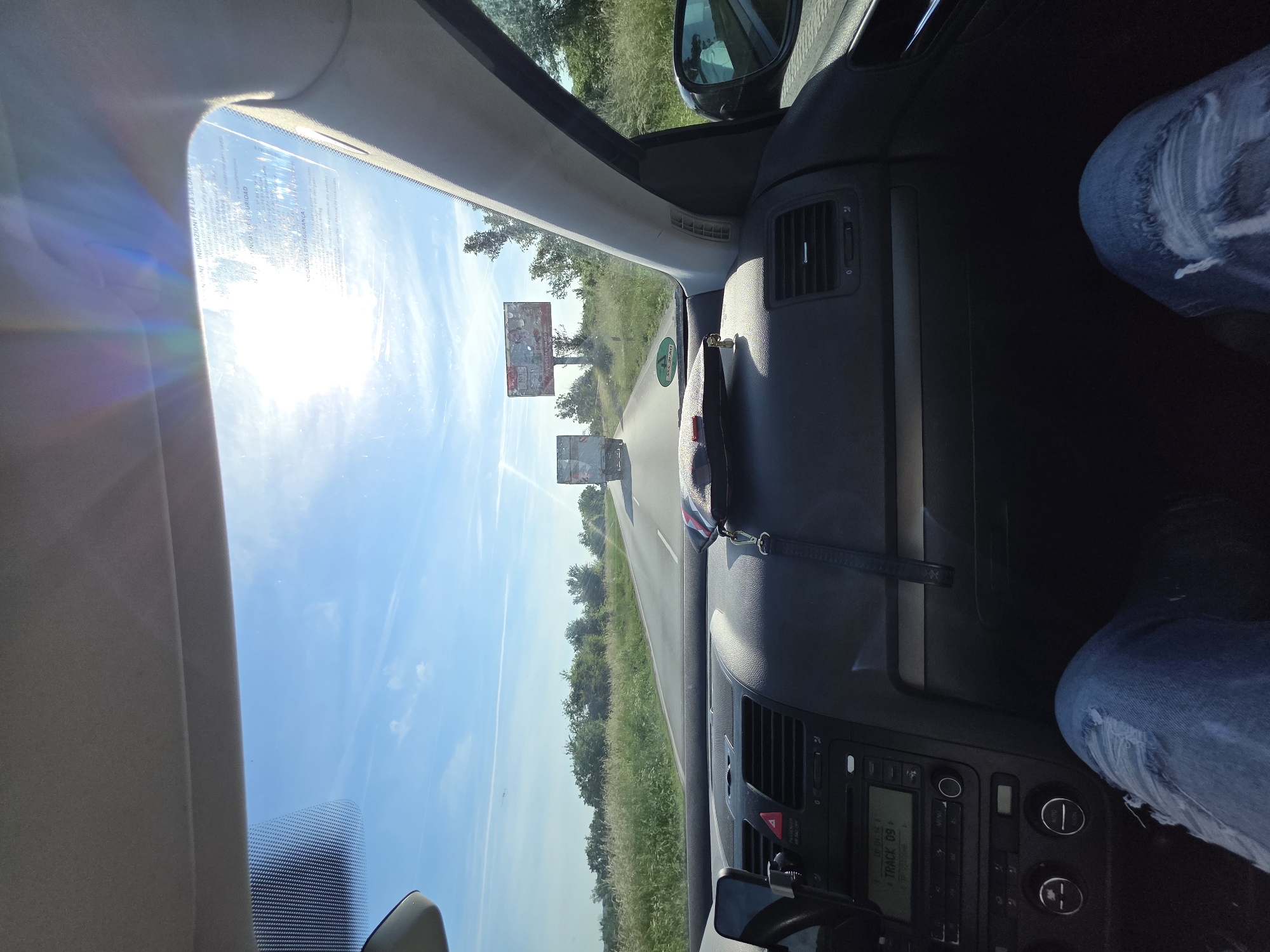 The image depicts a view from inside a car driving on a road in what appears to be a rural area near Novo Selo, Bulgaria. The sun is shining brightly through the windshield, causing glare. Two billboards are visible in the distance along the road. Greenery lines both sides of the road. Inside the car, part of the dashboard and the driver's leg are visible. The radio display shows "TRACK 09."