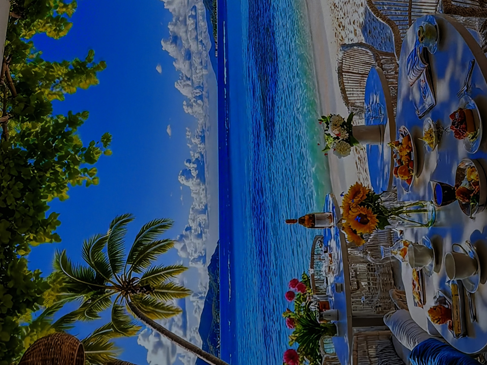 The image displays a vibrant and luxurious outdoor dining scene at a tropical beach resort, viewed from a perspective looking out towards the ocean. In the foreground, on the right side, multiple round and oval dining tables are elegantly set for a meal, likely breakfast or brunch. Each table features white place settings including plates, cups, glasses, and cutlery, along with various food items such as pastries, fresh fruit in bowls (berries, sliced fruit), and other dishes.
Decorative floral arrangements adorn the tables, with prominent bouquets of bright yellow sunflowers and arrangements of red and white flowers in clear vases. A bottle of what appears to be wine or another beverage is visible on one table. The chairs are made of wicker or rattan, complementing the natural, upscale ambiance.
The midground showcases a pristine white sandy beach leading to the calm, crystal-clear turquoise waters of the ocean near the shore, transitioning to a deeper blue further out. Gentle ripples are visible on the water's surface. In the distance, faint outlines of mountains or land masses can be seen across the expanse of the ocean, under a brilliant blue sky dotted with fluffy white clouds.
On the left side of the image, lush green tropical vegetation, including the prominent fronds of a palm tree and other leafy trees, frames the idyllic scene. The overall atmosphere conveys serenity, luxury, and natural beauty. The time of day is bright daylight, indicating clear and sunny weather. There is no visible text in the image. The visual evidence strongly indicates a tropical or subtropical coastal location.