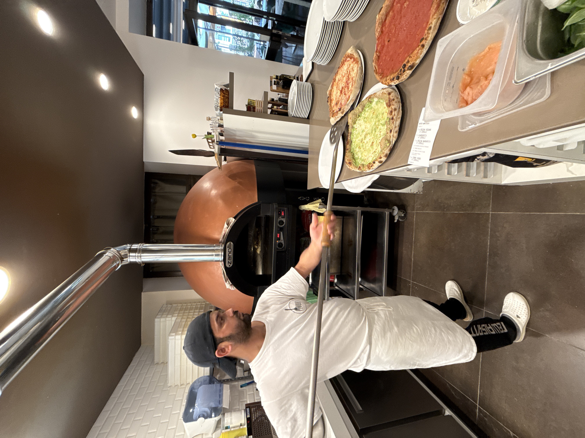 A man wearing a white t-shirt, white apron, black pants, and white crocs is standing in a pizzeria, holding a pizza peel. He is wearing a black baseball cap. In front of him, on a counter, are three pizzas: one with a green topping, one with a red tomato sauce topping, and another with a red tomato sauce topping. Behind him is a large, copper-domed pizza oven. The oven has a stainless steel flue pipe and a digital display showing "356". To the left of the oven, there are white stacked containers. To the right of the oven, on a shelf, are bottles of oil, a small vase with a yellow flower, and other items. Further to the right, there are stacks of white plates. The floor is tiled in a dark brown color. The walls are white and beige. The ceiling is dark brown with recessed lighting. The scene appears to be a commercial kitchen in a pizzeria located in Trento, Italy, given the context of the dish and the Italian context.