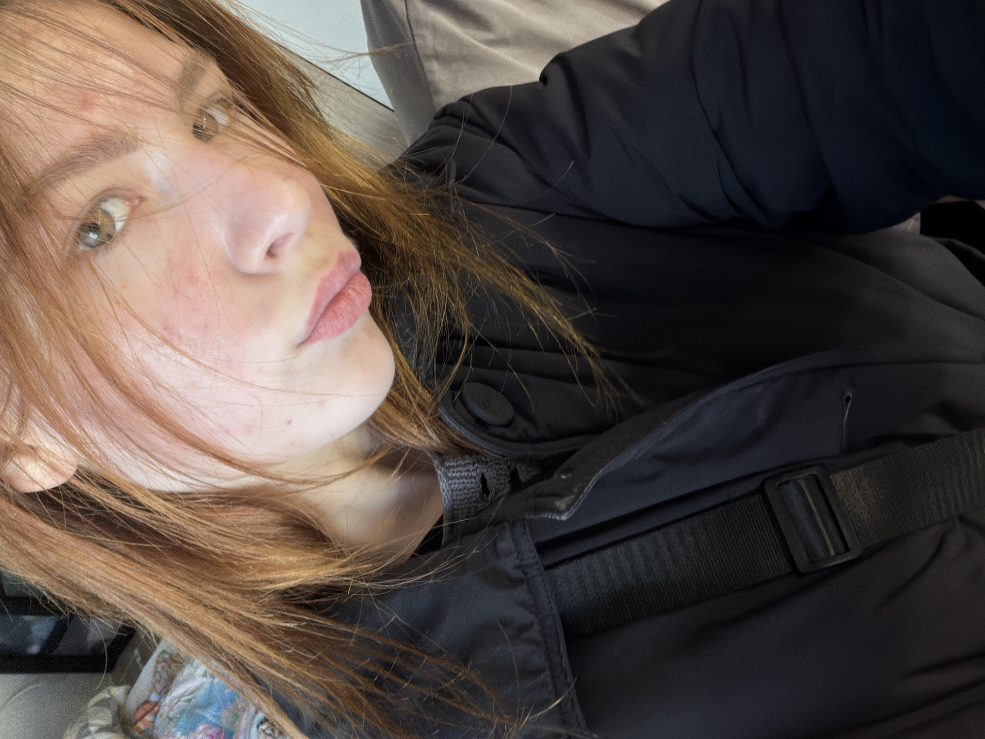 A young woman with light brown hair and fair skin is wearing a black jacket. The jacket has a strap across the chest. The woman is looking towards the camera with a slight tilt of her head. The background is out of focus, but suggests an indoor environment. The location in the city of Noviy, Russia is not determinable from the image.