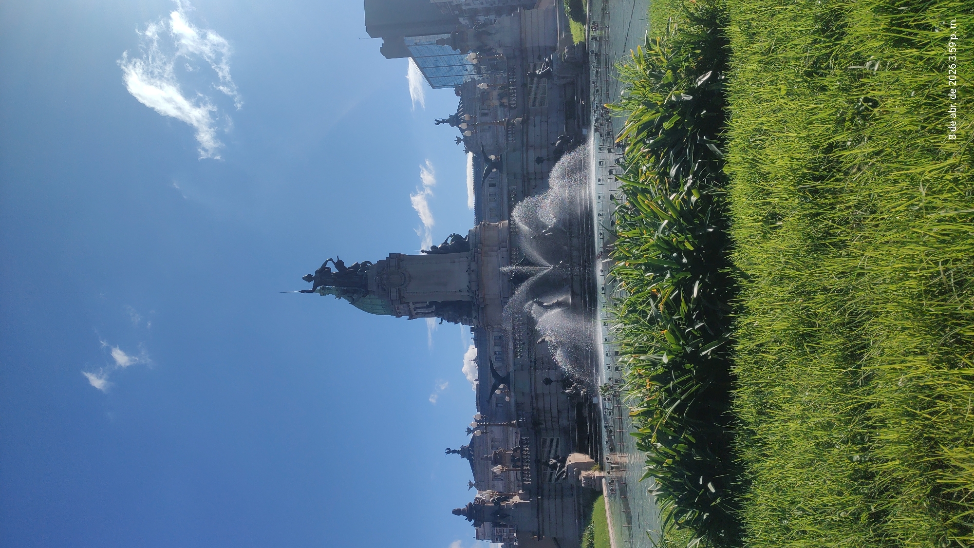 The image shows the Monument to the Carta Magna and Four Regions of Argentina, commonly known as the Monument of the Spanish, located in the city of Buenos Aires, Argentina. The monument is a large, ornate structure with sculptures and a fountain. The sky is clear and blue with some clouds. There is lush green grass and plants visible in the foreground.