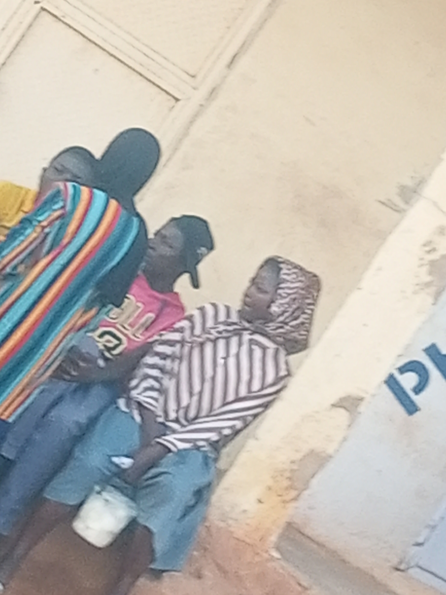 In an outdoor setting, likely a street or public area in Gombe, Nigeria, four individuals are visible. Three individuals are in the foreground, facing away from the camera or to the side. The person on the far left is wearing a horizontally striped shirt in shades of blue, orange, and yellow, with a dark garment underneath. To their right, another person wears a pink t-shirt with white lettering and a black cap. A third person, positioned further right and closer to the camera, is wearing a vertically striped shirt in black and white and a headscarf patterned with black and white swirls. This individual appears to be holding or interacting with a white object. The fourth individual is partially visible behind the first two. The background consists of a light-colored wall, possibly part of a building, with a diagonal shadow or beam. A portion of a light blue sign with dark blue lettering, appearing to be the letter "P", is visible on the right side of the frame. The ground beneath the individuals is a mix of dirt and possibly a rough surface.