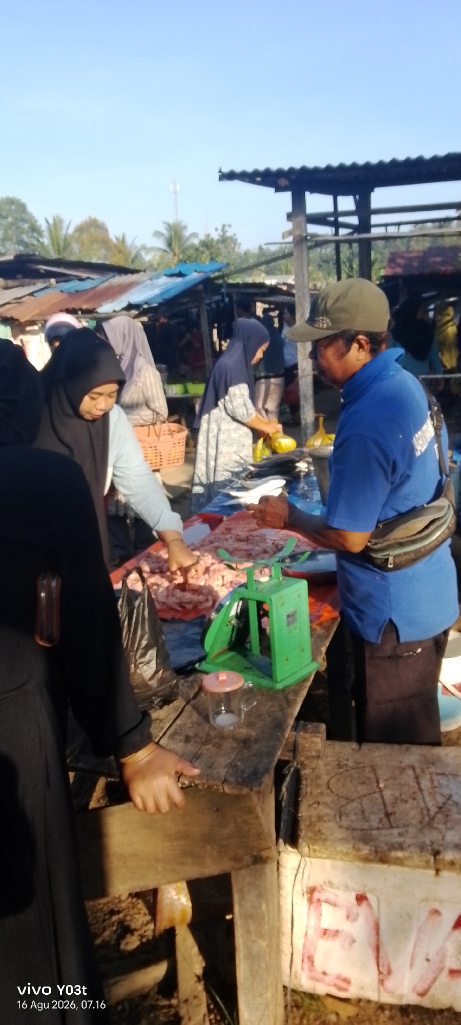The image depicts a bustling outdoor market scene in Palosilae, Indonesia. A man wearing a blue shirt and a cap is operating a green scale at a stall. In front of him, a woman in a black hijab is attending to a customer. Another woman, also wearing a hijab, is visible in the background, seemingly engaged in her own transactions. The stall is laden with various goods, including what appears to be raw meat or fish, arranged on a blue tarp. A black plastic bag and a small glass container are also present on the wooden surface. The market appears to be set up under makeshift shelters with corrugated tin roofs, indicative of a rural or semi-urban setting. The lighting suggests it is daytime, possibly late afternoon given the warm tones. The bottom left corner of the image displays a timestamp: "vivo Y03t 16 Agu 2026, 07.16".
