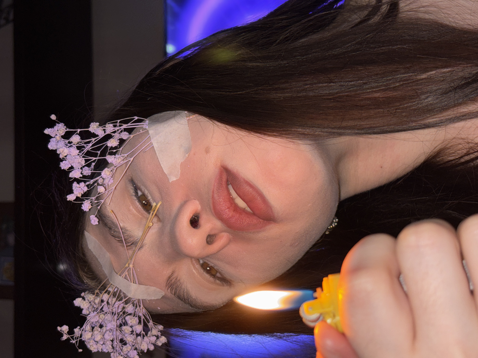 This image is a close-up, angled portrait featuring a person, likely a young woman, with her head tilted, looking slightly upwards. Her long, dark brown hair frames her face. Delicate sprigs of light purple dried flowers are adorned on her face and hair, held in place by strips of light-colored adhesive tape on her forehead and cheek. Her lips are slightly parted, and her eyes are open, reflecting a neutral or contemplative expression.
In the foreground, a hand holds a yellow plastic lighter, producing a bright orange flame. This flame acts as the primary light source, casting warm, glowing illumination onto the person's face and hair. The background is dark, but a distinct blue-purple light emanates from an unseen source, possibly a television screen or decorative lighting.
The setting appears to be indoors, likely in a dimly lit room, suggesting it is night or evening. The overall composition is intimate and somewhat artistic, focusing on the interplay of light, the person's features, and the unusual adornments. The image provides no specific location cues beyond the context of Perm, Russia. There is no visible text in the image.