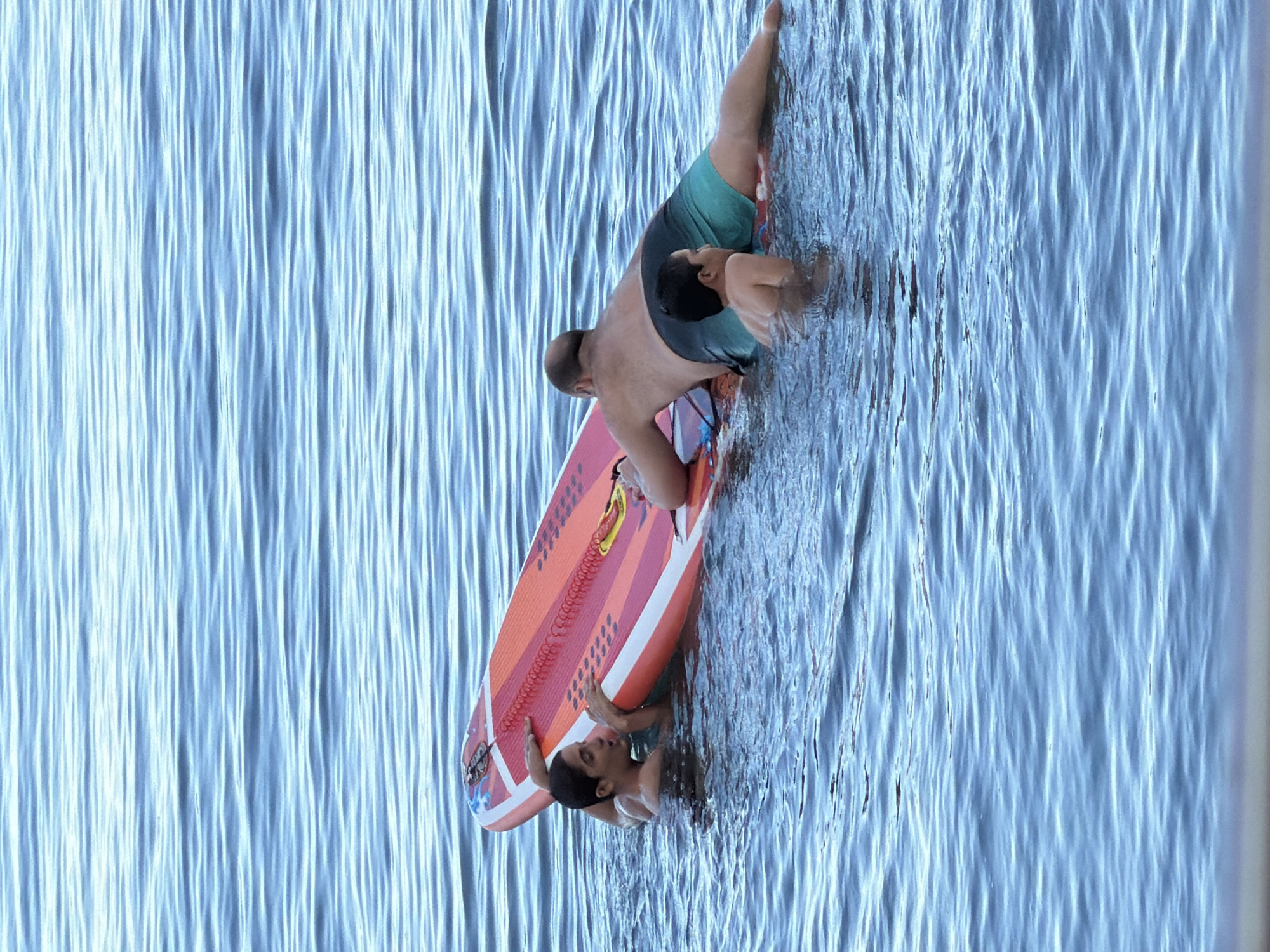 The image shows two individuals in the water, seemingly engaged in an activity involving a surfboard. One person, wearing teal swim trunks, is partially submerged, with their upper body visible. They appear to be holding onto a surfboard, which is predominantly orange and red with white accents and black lettering. A second person, a child, is also in the water, positioned partially beneath the surfboard and against the person in swim trunks. The water is a vibrant blue, with noticeable ripples and waves, suggesting movement. The scene appears to be set outdoors, with ample natural light, implying it could be daytime. The overall mood seems relaxed and possibly playful, given the water activity and the positioning of the individuals. The presence of the surfboard and the water suggests a recreational setting, likely a beach or a lake. There are no distinct location cues other than the given context of Partenit, Ukraine. No text is clearly legible on the surfboard or elsewhere in the image.