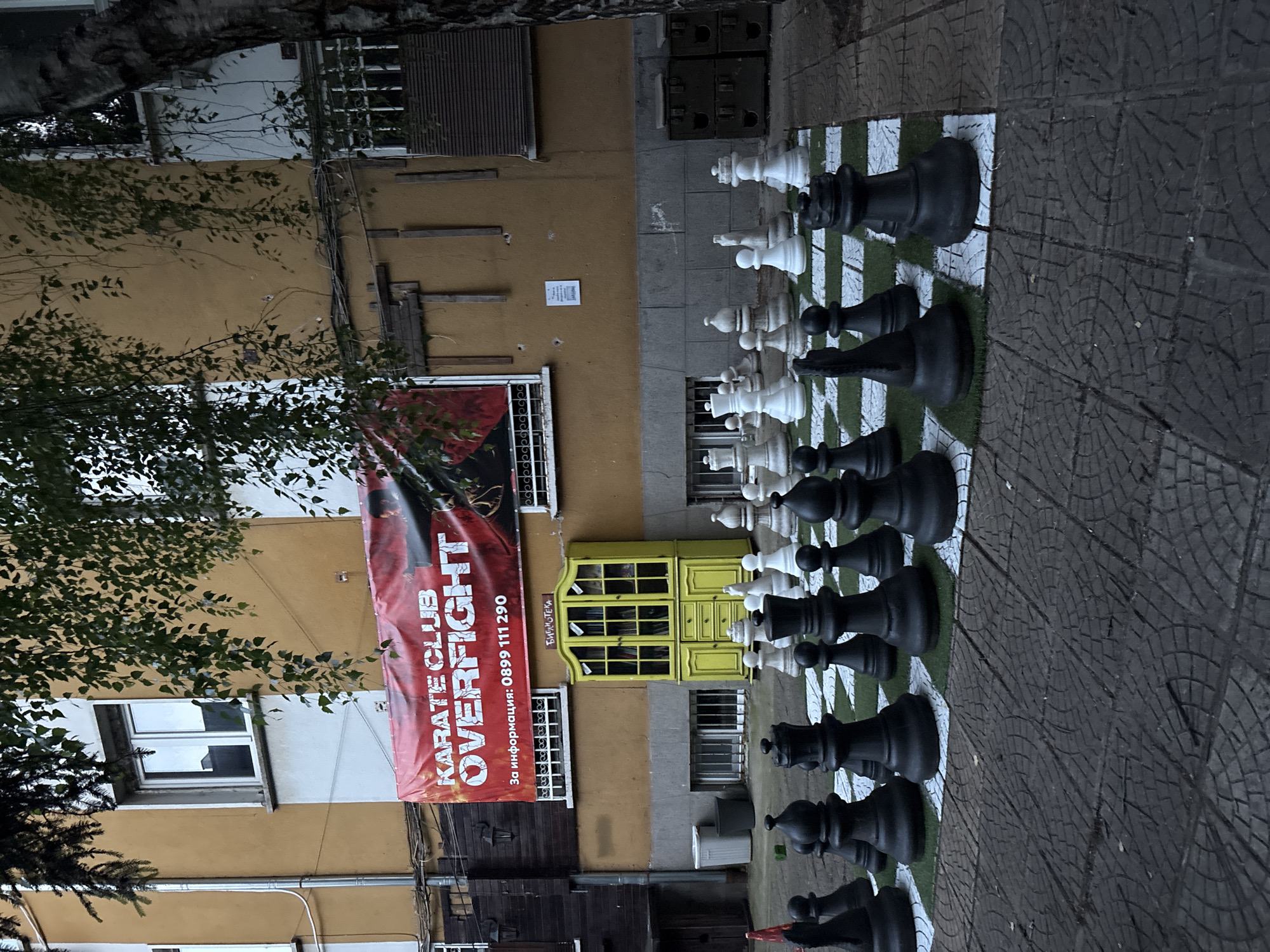 In an outdoor courtyard in Sofia, Bulgaria, a giant chess set is arranged on a checkered green and white mat. The chess pieces, both black and white, are large and made of plastic. Dominating the background is a building with a mustard-yellow facade, featuring several windows and a red banner that reads "KARATE CLUB OVERFIGHT". Below the banner, a bright yellow cabinet with glass doors, labeled "Библиотека" (Library), stands against the building. The courtyard pavement is made of grey interlocking tiles. Several trees with green foliage hang over the scene, obscuring parts of the building and sky. The lighting suggests it is daytime, possibly overcast or late afternoon, with some shadows cast by the chess pieces and trees.