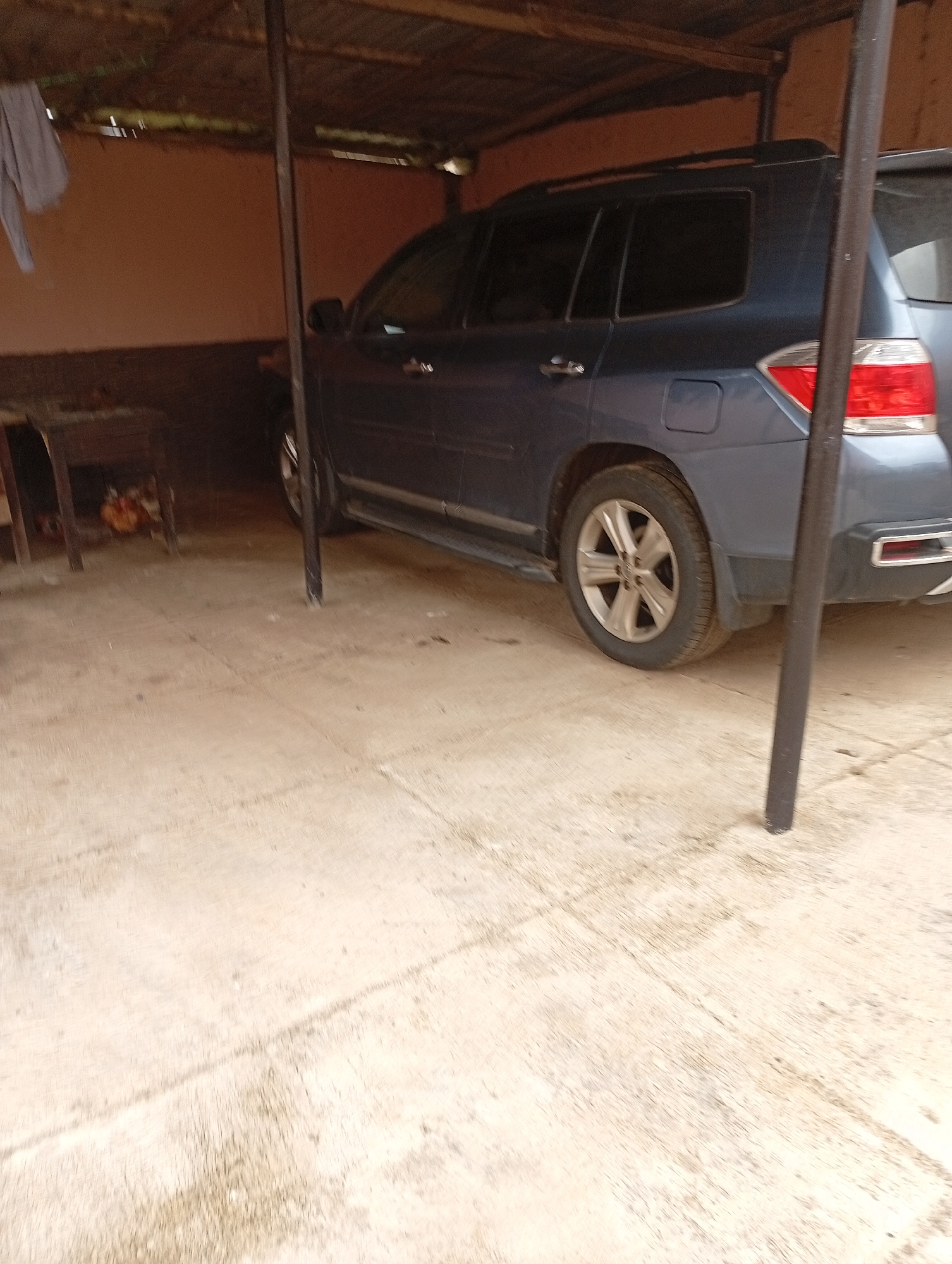 The image depicts a dark blue SUV parked under a makeshift shelter. The shelter appears to be constructed with corrugated metal roofing supported by wooden beams and metal poles. To the left of the SUV, a small wooden table with items on it is visible. The ground is concrete, and the walls of the shelter are a reddish-brown color. The scene is outdoors, suggesting a parking area or yard. The location is identified as Rafin Par, Nigeria. There are no people visible in the image, and the SUV is stationary. The lighting suggests it might be daytime, possibly overcast.