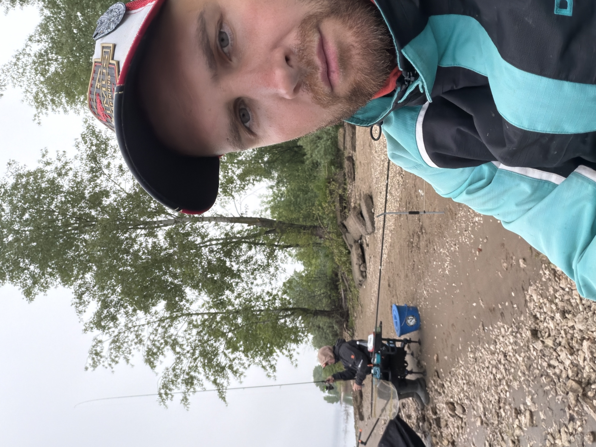 This vertically oriented image is a selfie taken by a young man, rotated ninety degrees clockwise. He has a light beard and is wearing a red and white baseball cap with a black brim, on which the word "SPINNING" and part of "ULTIMATE" are visible as a gold and red logo. He is also wearing a jacket with teal/light blue and dark blue/black sections. His expression is neutral as he looks at the camera.
The setting is an outdoor fishing spot, likely a river or lake bank in Noviy, Russia, under an overcast, grey sky. In the mid-ground, another person is actively fishing, seated on a specialized fishing chair or station with a rod extended over the water. Several fishing rods are propped up near the water's edge, and a blue bucket is visible on the ground. The bank itself is composed of mud and a significant amount of small, light-colored gravel and stones. Trees with green foliage line the far bank or shoreline, suggesting a natural, secluded environment. The overall scene depicts a fishing activity on a cloudy day.