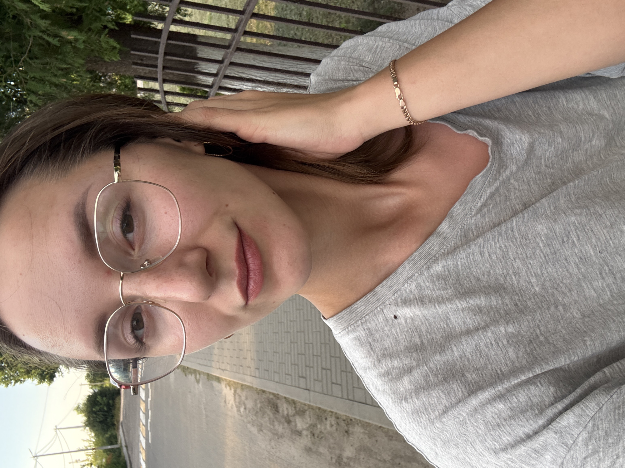 This is a vertical, close-up, selfie-style photograph of a young woman with brown hair and glasses. She is wearing a light gray t-shirt. Her left hand is raised to her head, touching her hair. A thin gold bracelet is visible on her right wrist. She is wearing light-rimmed glasses and has light pink lipstick on her lips. The background shows a blurry outdoor scene with trees, a fence, and a paved walkway. The lighting suggests it is daytime, likely late afternoon or early evening, given the warm tones. The image has a casual and relaxed feel, possibly taken while sitting or standing outdoors. The location is identified as Zhodzina, Belarus. There is no visible text in the image.