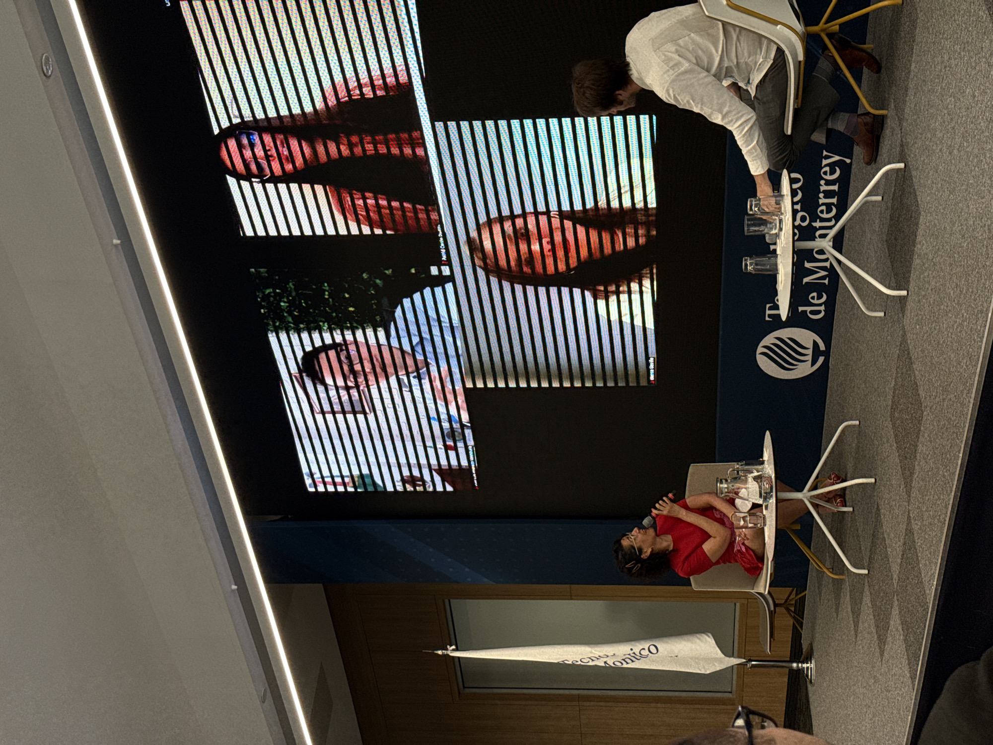 An indoor event is depicted, likely within a conference room or auditorium at the Tecnológico de Monterrey, Campus Ciudad de México. The scene is dominated by a large, horizontal LED screen displaying four video conference feeds, positioned above a dark blue wall featuring the "Tecnológico de Monterrey" logo and text.
Two individuals are seated on a carpeted stage area in the foreground. On the left, a woman with dark, curly hair and glasses, wearing a red short-sleeved top and a patterned skirt, holds a black microphone to her mouth, indicating she is speaking. She is seated in a light-colored armchair with a white frame, beside a small white round table holding a glass pitcher of water, a napkin, and two glasses. To the left of her, a white flag with vertical text, likely "Tecnológico de Monterrey", stands on a pole.
On the right, a man is seated in a light-colored armchair with a yellow frame. He wears a light-colored long-sleeved shirt, dark pants, striped socks, and brown dress shoes. His back is mostly to the viewer, but his head is turned towards the screen or the woman, suggesting he is observing. A second small white round table sits in front of him, holding two glasses.
The large screen displays four distinct video frames. The top-left frame shows a man with glasses. The top-right frame shows a woman with long dark hair and glasses; the name "Astrid Carola Duarte" is partially visible below her feed. The bottom-left frame shows a woman with long dark hair, and the name "Marisa Gaviria" is visible below her feed. The bottom-right frame appears dark or empty. Horizontal scan lines are visible across the entire LED screen. The ceiling above features a long, recessed linear light fixture. The background includes wooden wall paneling and a dark blue vertical column.